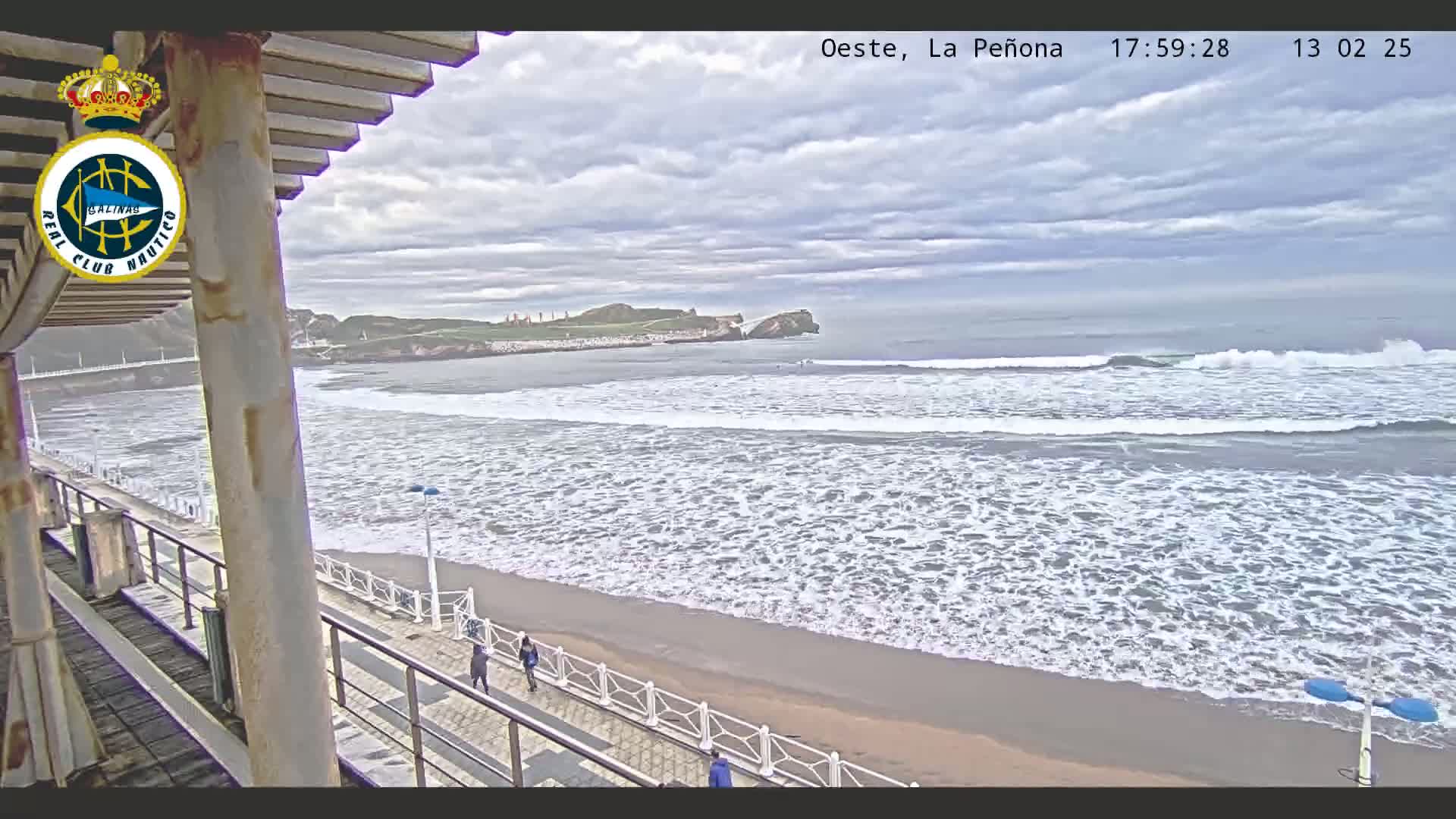 A beach scene shows a partly cloudy sky over a sandy beach with gentle waves rolling in; a few people stroll along a paved walkway next to the water.