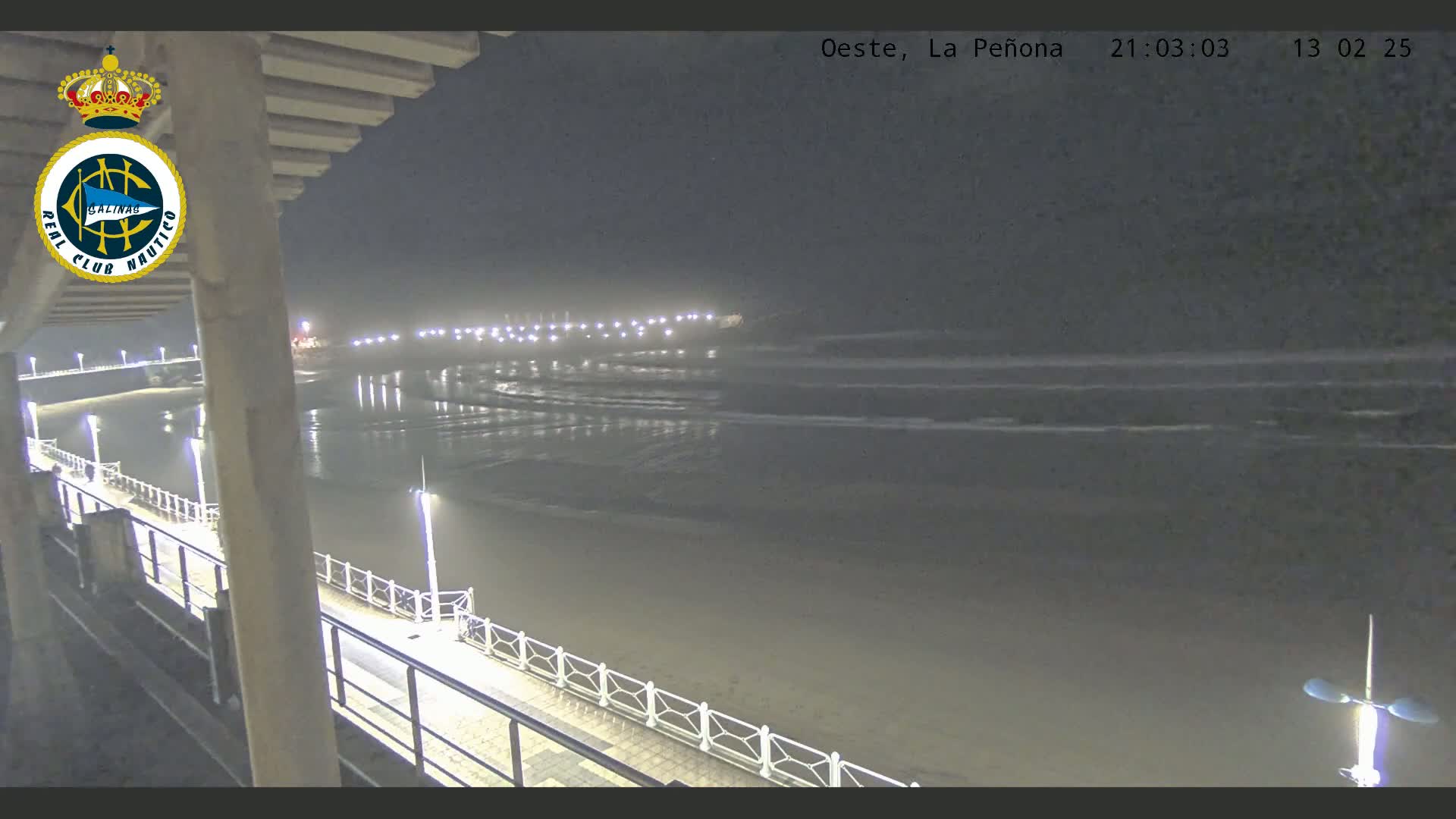 A nighttime view from an elevated position shows a dark, gently lapping ocean with a long, lit walkway along a beach.