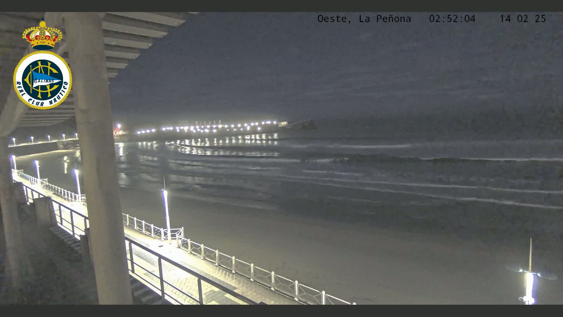 A nighttime view from an elevated position shows a beach with gently lapping waves under a dark, mostly clear sky, and a pier extending into the water, illuminated by lights.