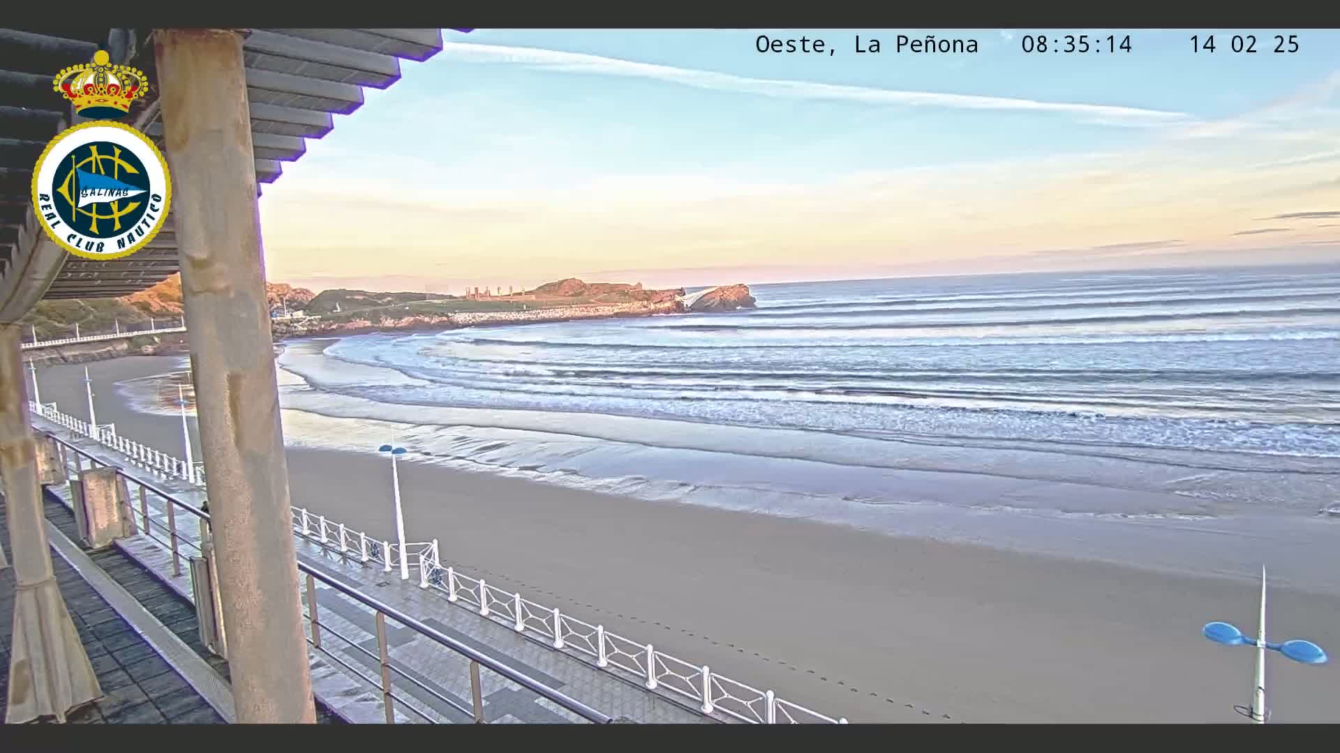 A sandy beach with gentle waves under a clear, light-pink sunrise sky is viewed from an elevated position on a boardwalk.