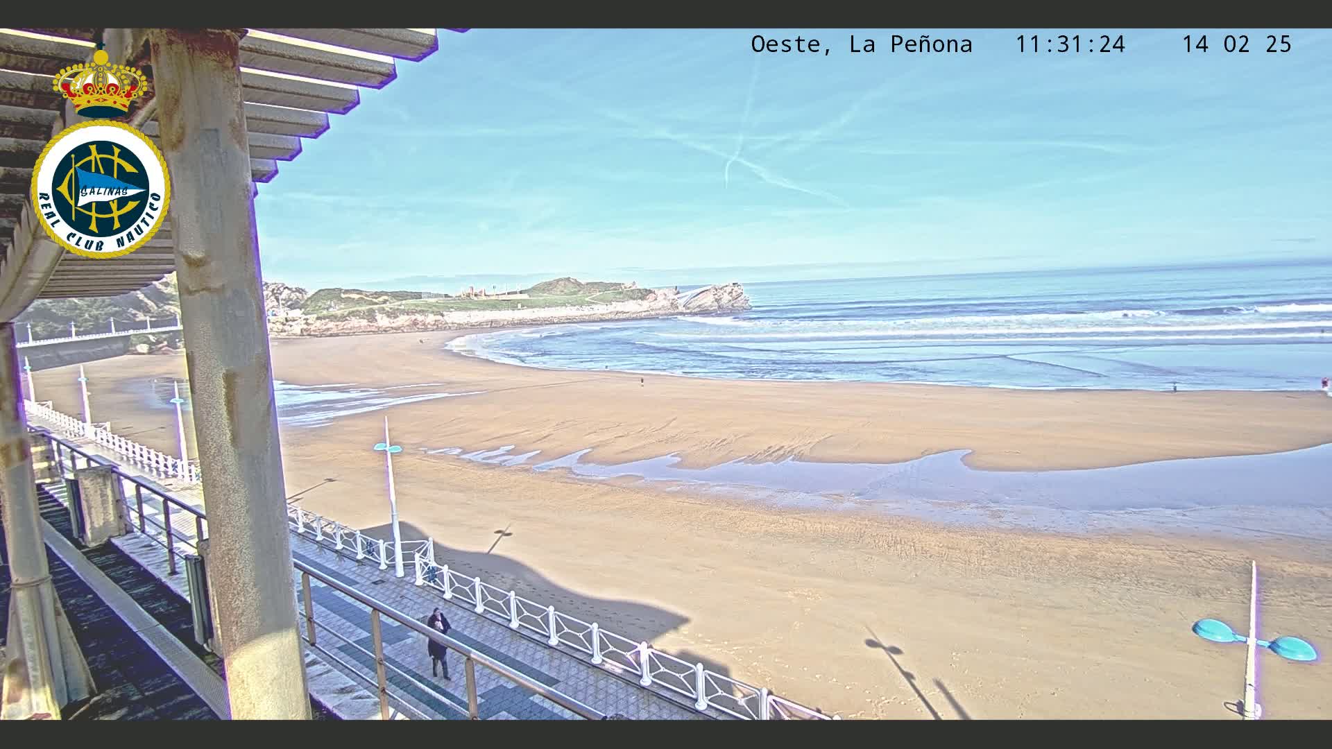 A sandy beach stretches to a calm ocean under a clear blue sky, with a person walking along a boardwalk that overlooks the scene.