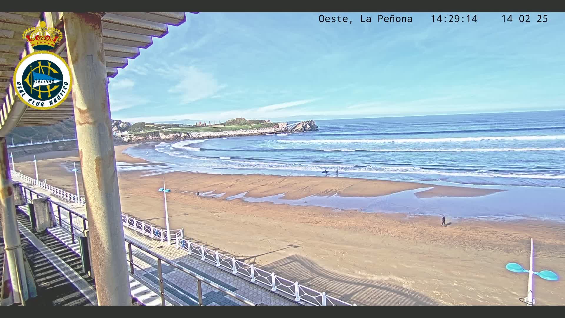 A sandy beach with gentle waves under a clear blue sky is seen from an elevated viewpoint, overlooking a boardwalk and a rocky outcrop in the distance.