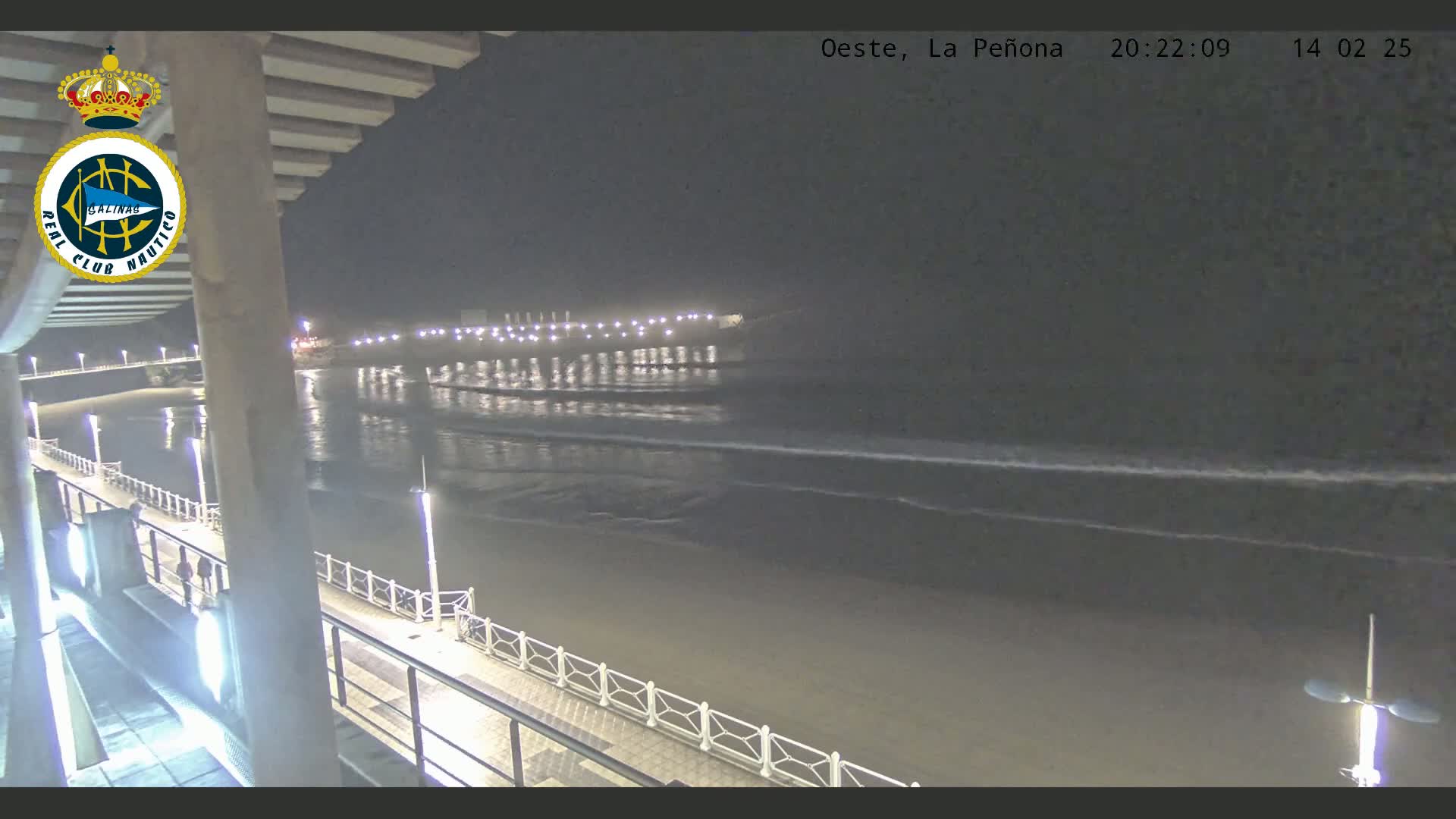 A nighttime view from a pier shows a dark sandy beach with gentle waves lapping the shore under a clear, dark sky.