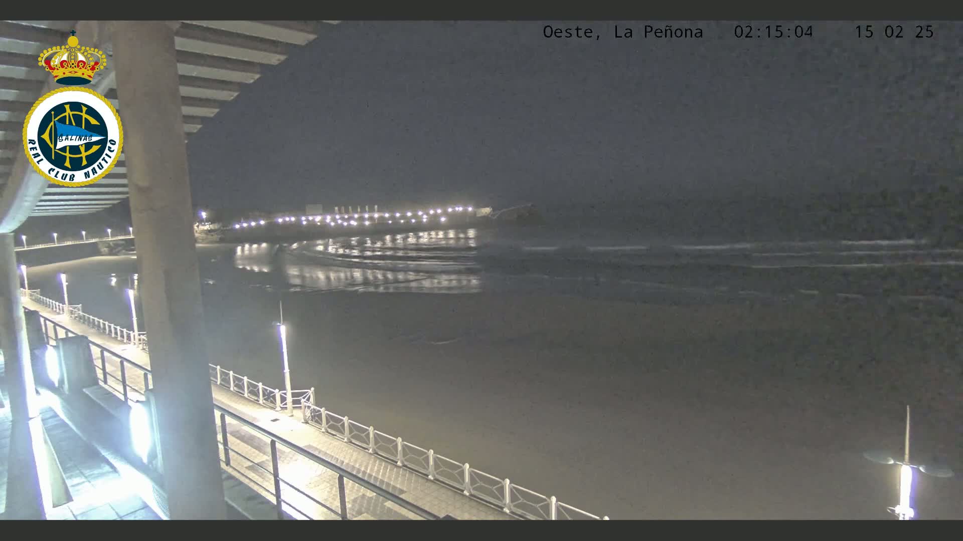 A nighttime view of a calm ocean with small waves lapping a shore, seen from a pier with lights illuminating the walkway.