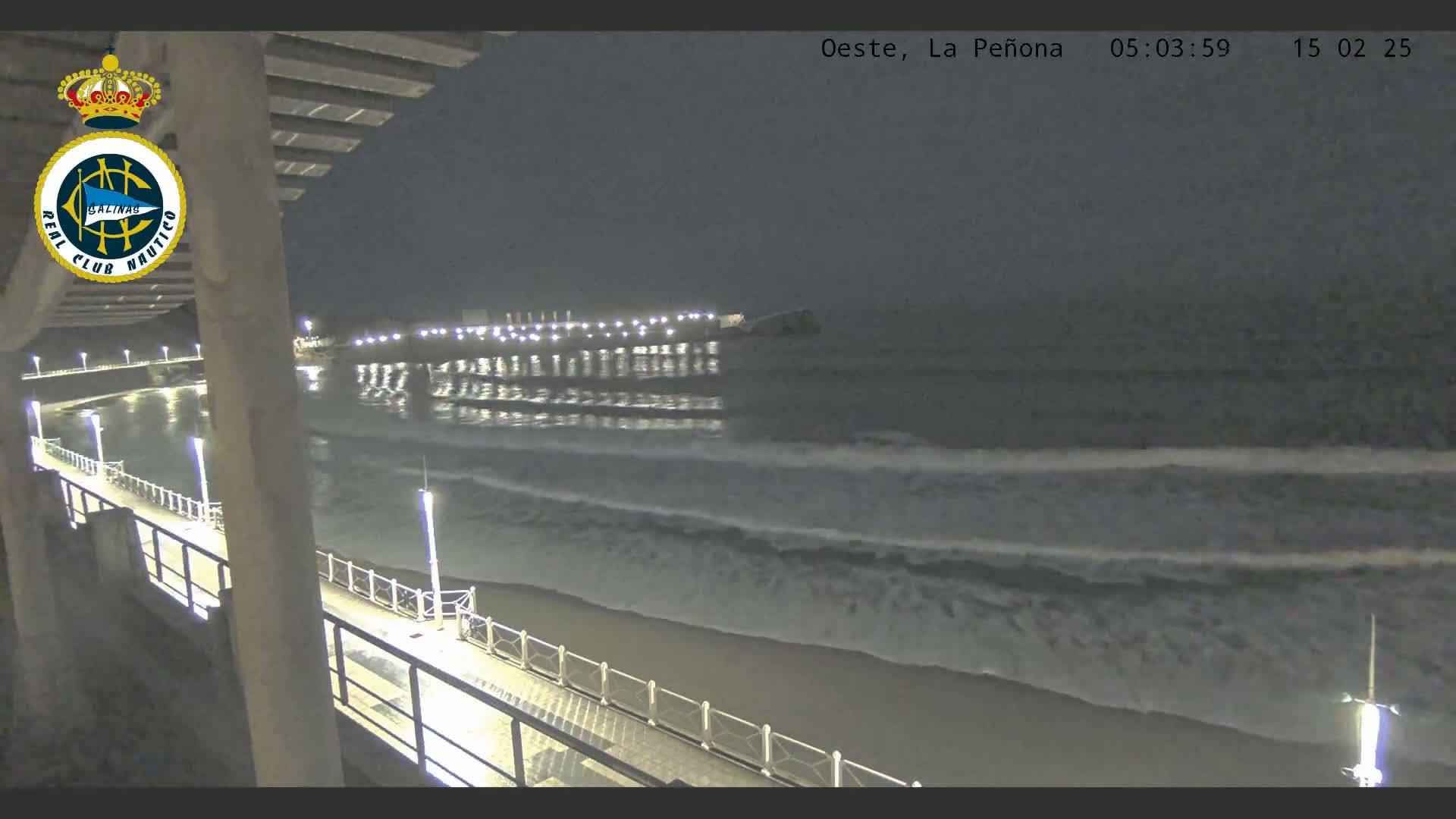 A nighttime view from a pier shows a calm ocean with small waves gently lapping the shore, illuminated by lights along the pier and a building in the distance.