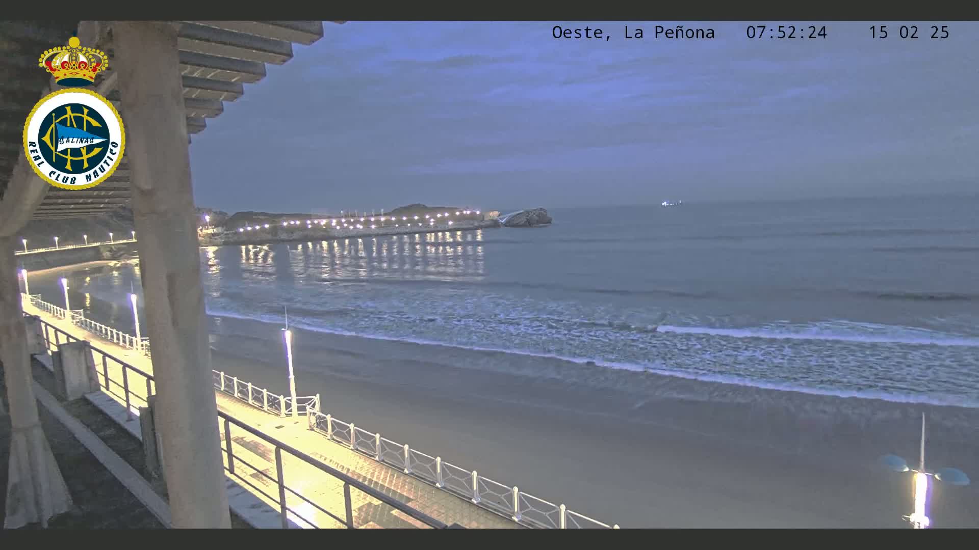 A nighttime beach scene shows a sandy shore with gentle waves lapping the sand under a twilight sky, with a boardwalk and lights visible along the beachfront.