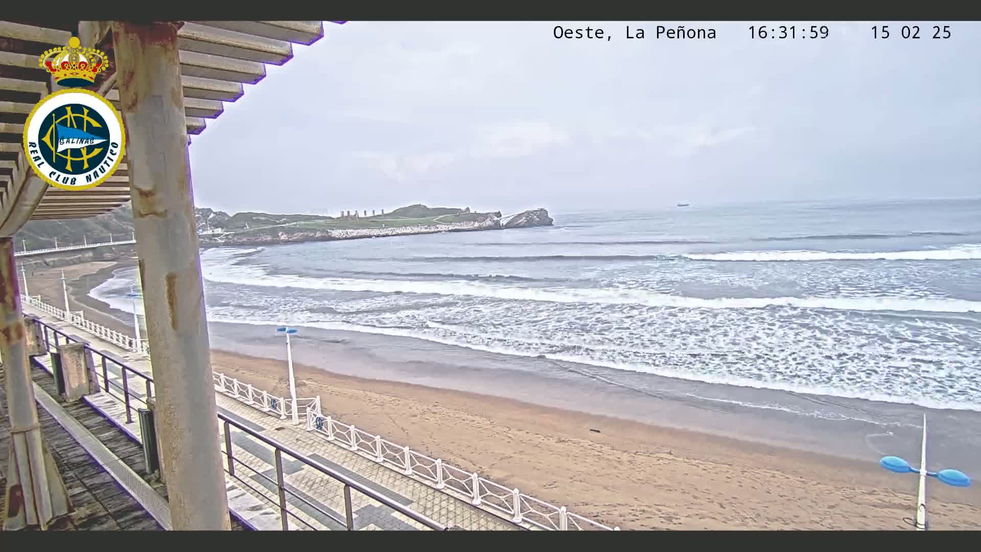 A sandy beach with gentle waves under an overcast sky is viewed from an elevated position, showing a boardwalk and a distant rocky outcrop.