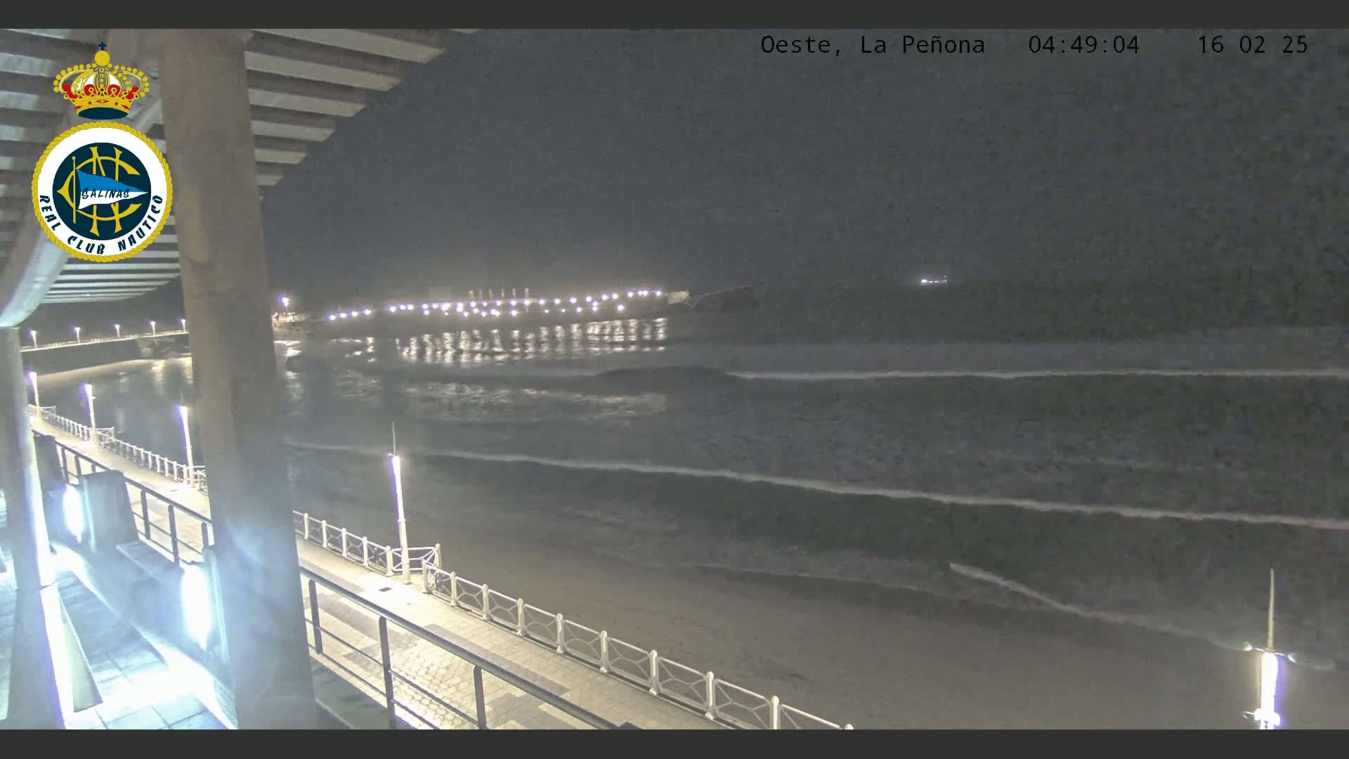 A nighttime view of a beach with gentle waves lapping the shore, a pier extending into the water, and a walkway lined with lights.