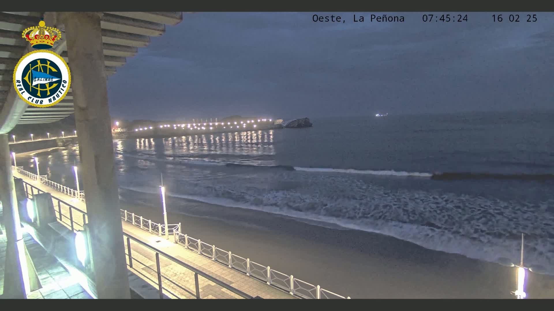 A nighttime view from an elevated position shows a long pier extending into a dark ocean with small waves lapping a sandy beach, all lit by artificial lights.