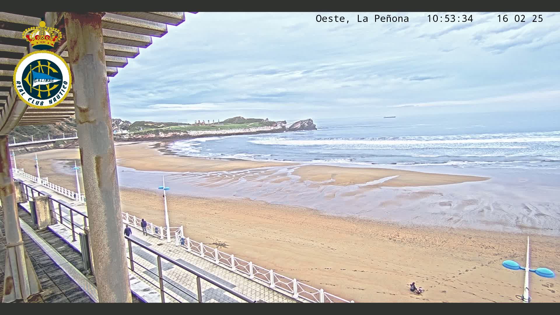 A sandy beach with gentle waves under a cloudy sky is viewed from a partially covered walkway.
