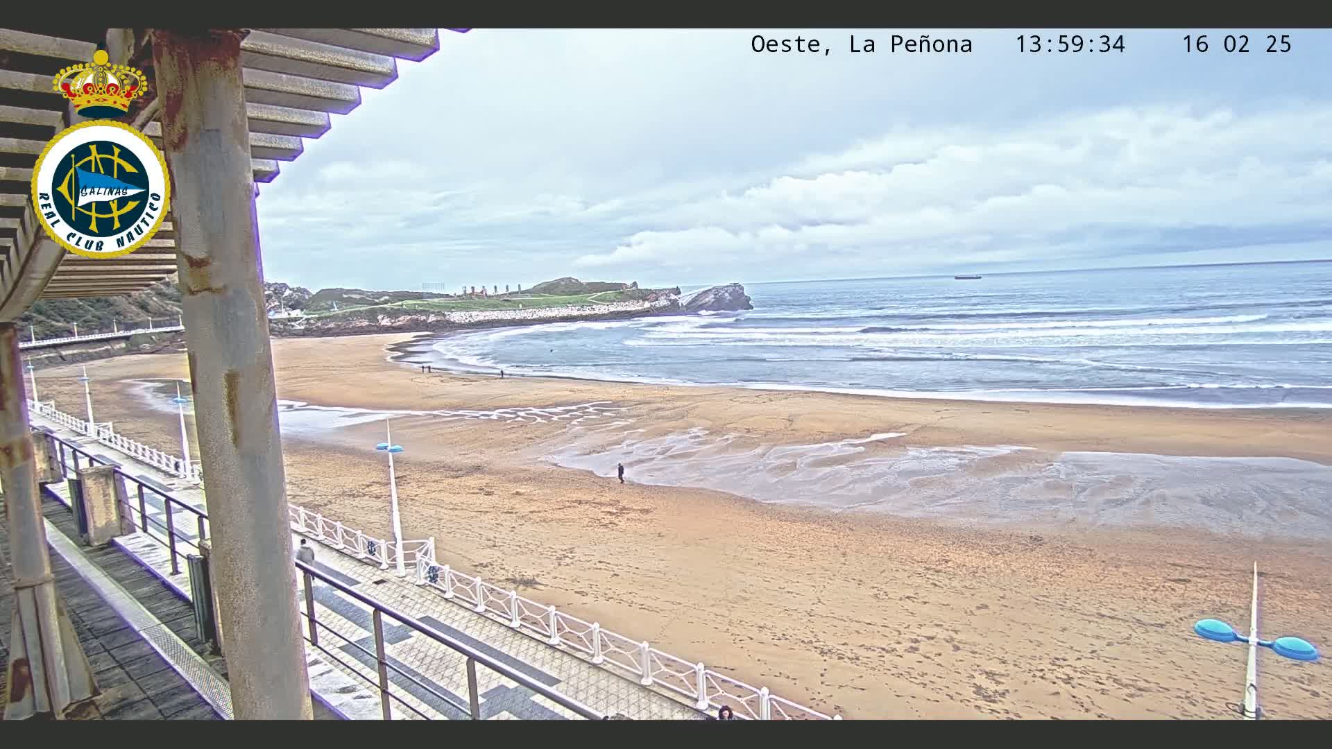 A sandy beach with small waves under a cloudy sky is viewed from a covered balcony.