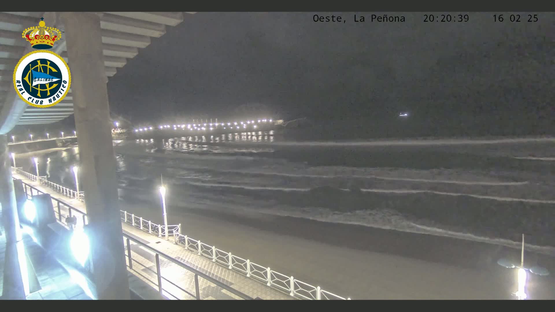 A nighttime view of a beach with gentle waves lapping the shore, illuminated by lights along a pier and boardwalk.