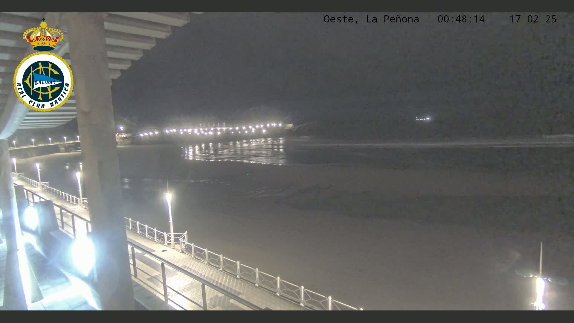 A nighttime view from a pier shows a dark beach and calm ocean water under a clear sky, with lights illuminating the pier and the shoreline in the distance.