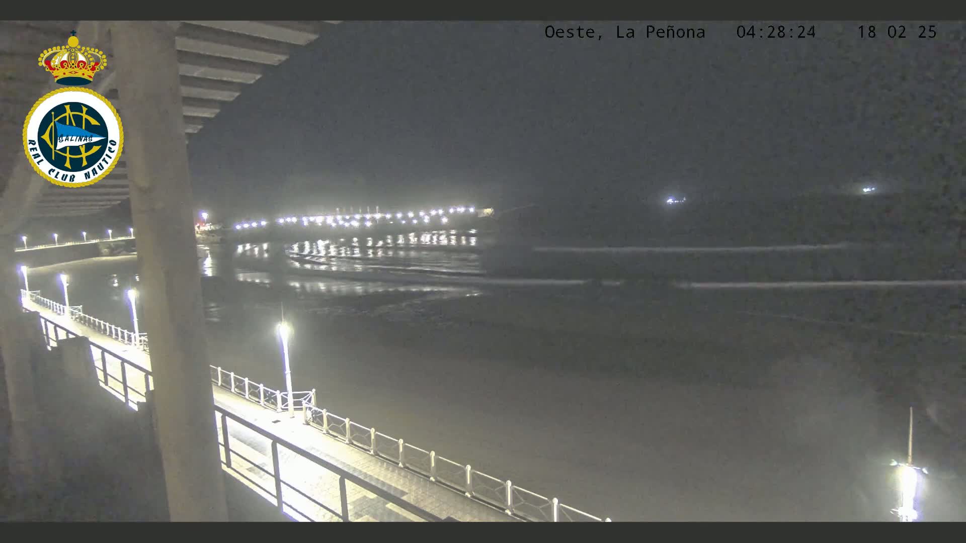 A nighttime view from a pier shows a calm, dark ocean reflecting lights from a distant shore.