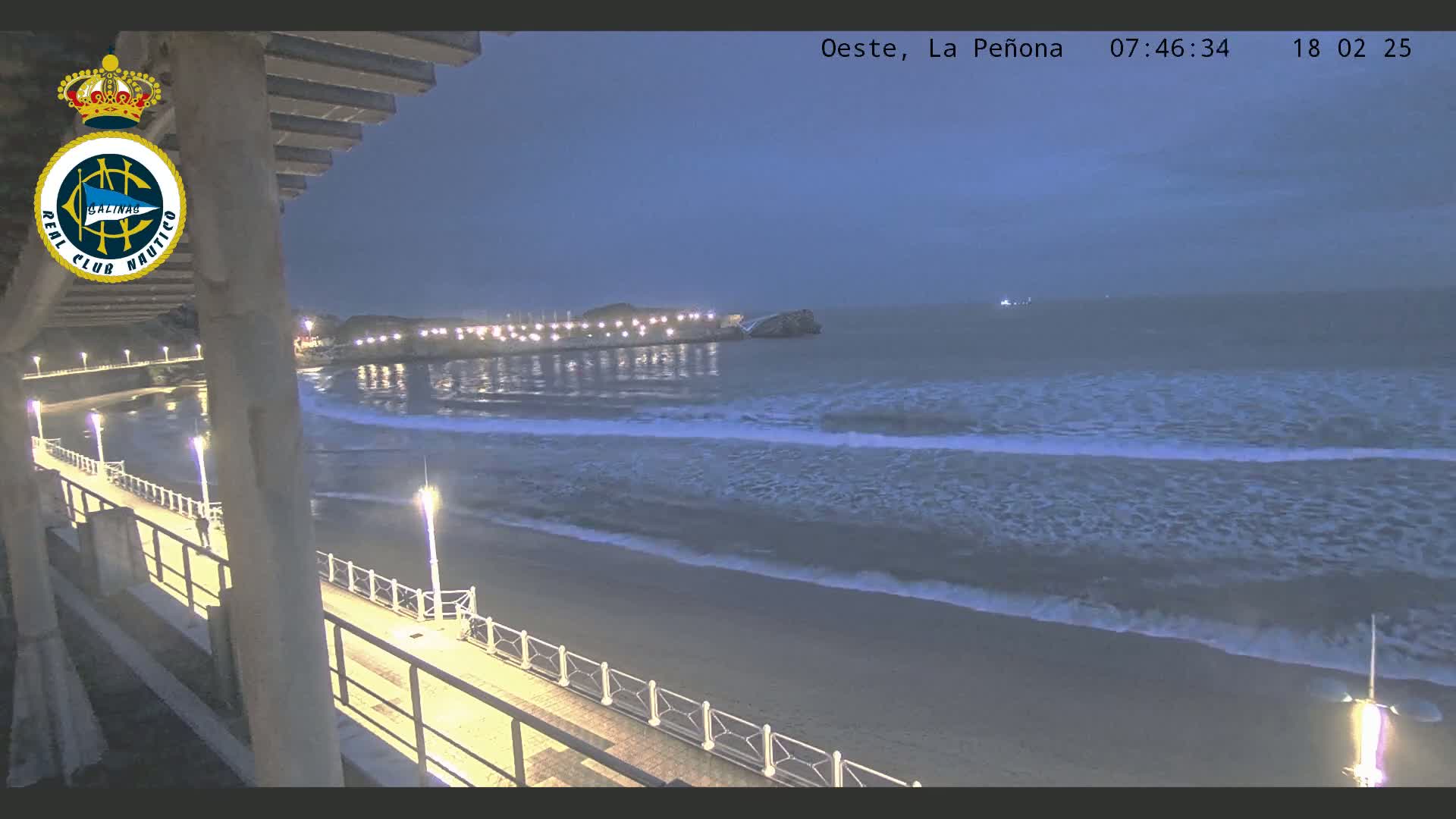 A nighttime view of a beach with gentle waves rolling onto the shore, illuminated by streetlights along a boardwalk, under a twilight sky.