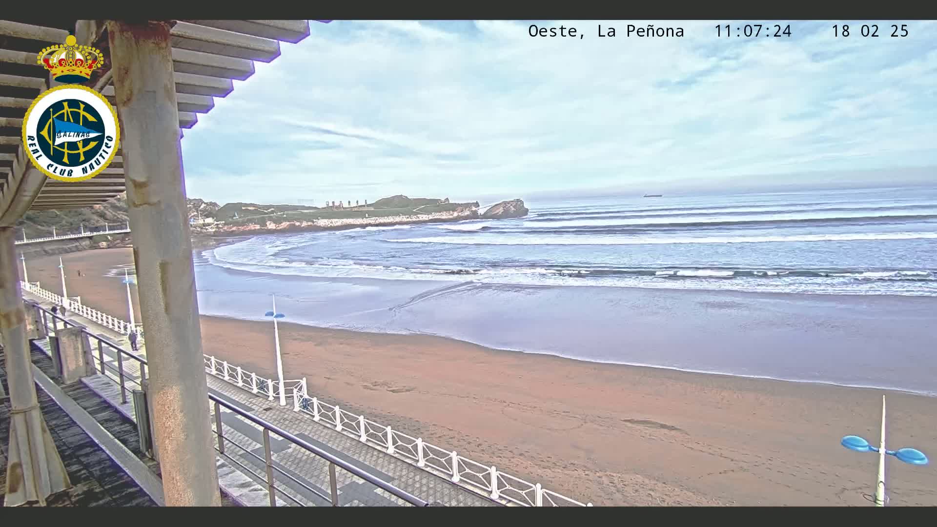 A sandy beach with gentle waves under a partly cloudy sky is viewed from an elevated position, showing a boardwalk and a distant rocky outcrop.