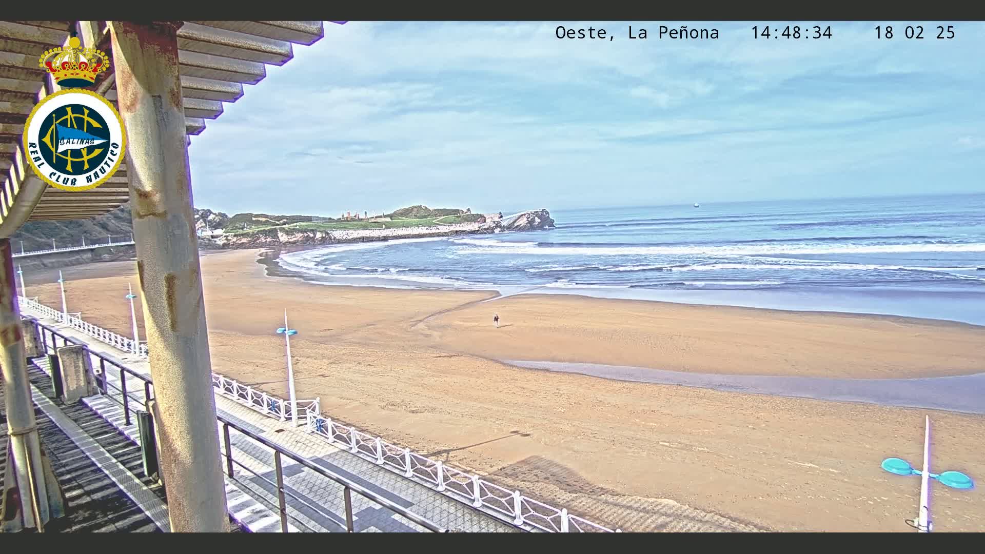 A sandy beach with gentle waves under a partly cloudy sky is viewed from a walkway with a railing.