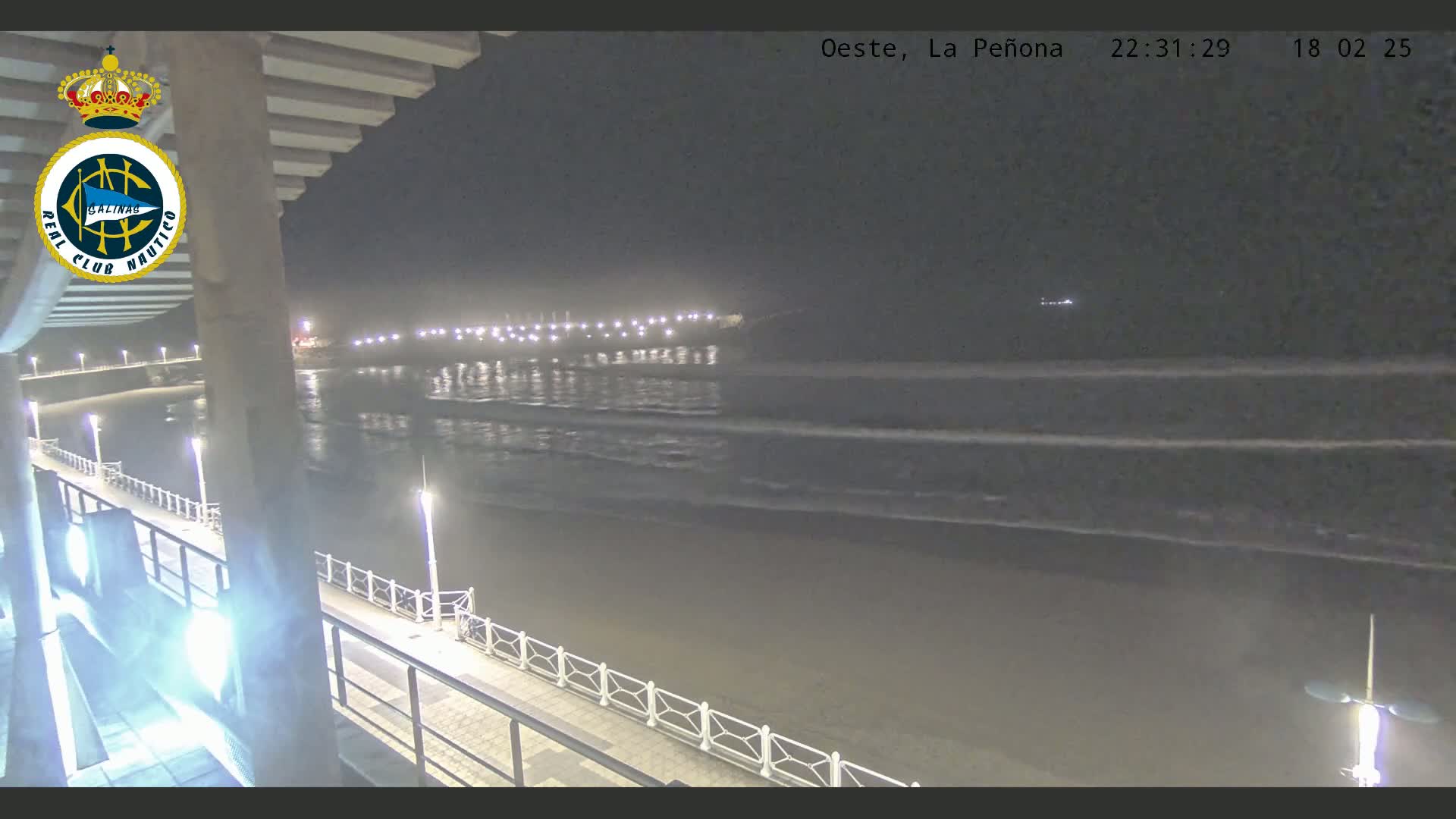 A nighttime view of a beach with gently rolling waves, a boardwalk with lights, and a pier in the distance.