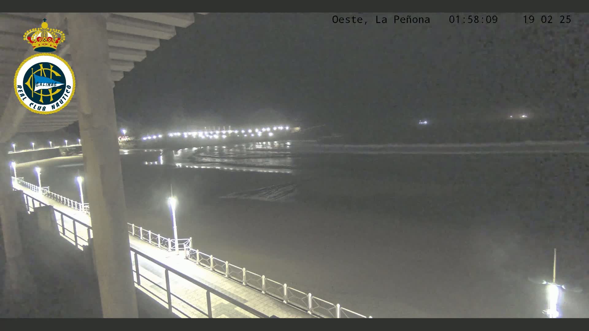 A nighttime view of a pier extending over a dark ocean with gentle waves, illuminated by lights along the pier and a distant shoreline.