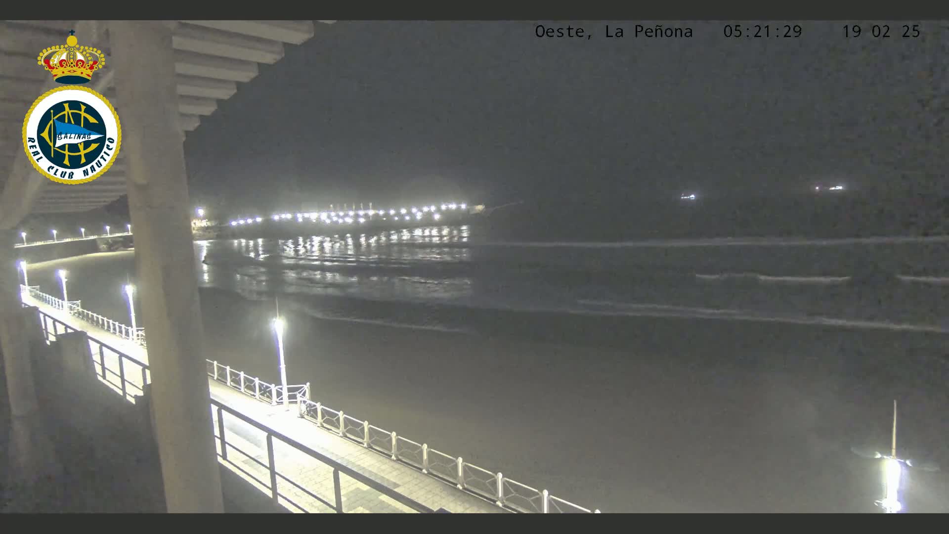 A nighttime view from a pier shows a dark ocean with gentle waves, a shoreline with lights, and a walkway lit by lamps.