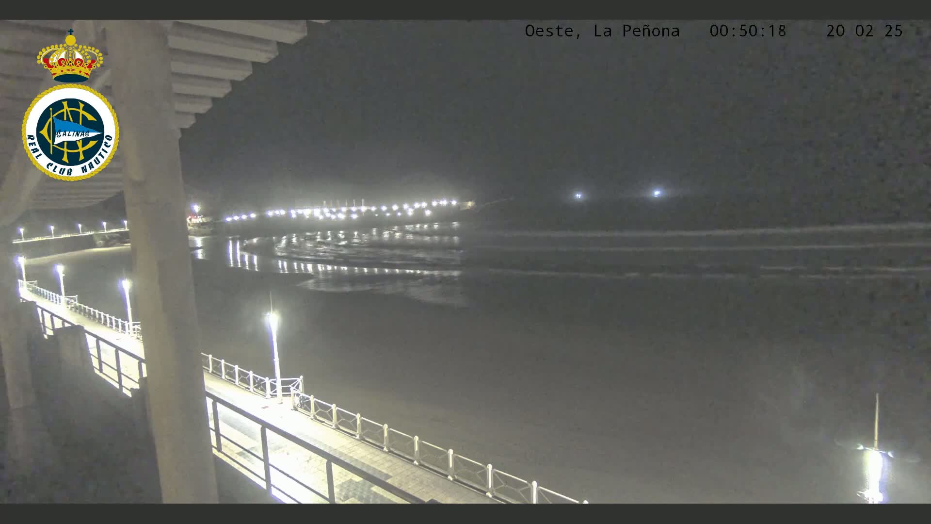 A nighttime view from a pier shows a dark beach and calm ocean with gentle waves under a clear sky, illuminated by lights along the pier and in the distance.