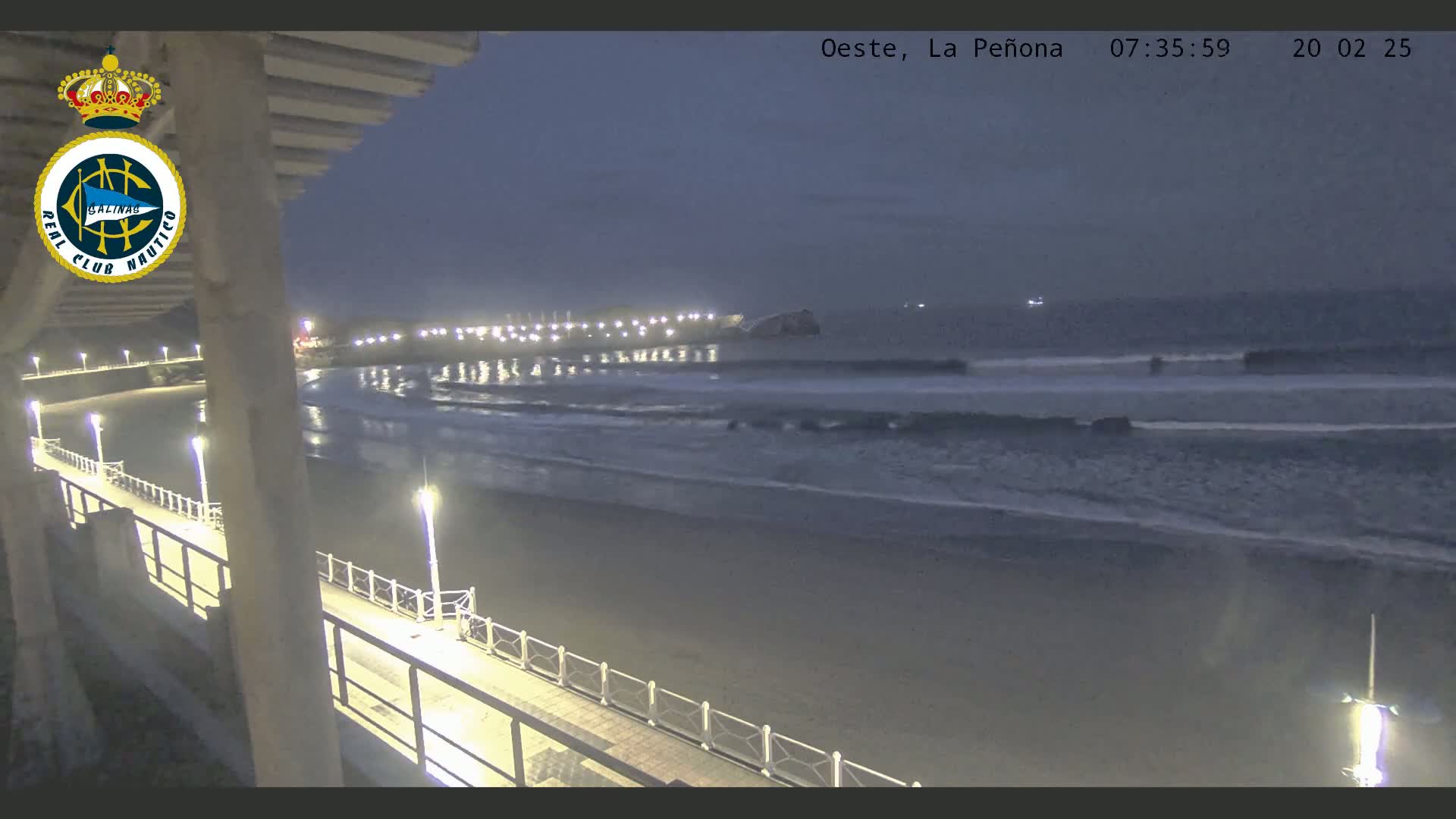 A nighttime view from a pier shows a dark beach with gently lapping waves under a dusky sky.