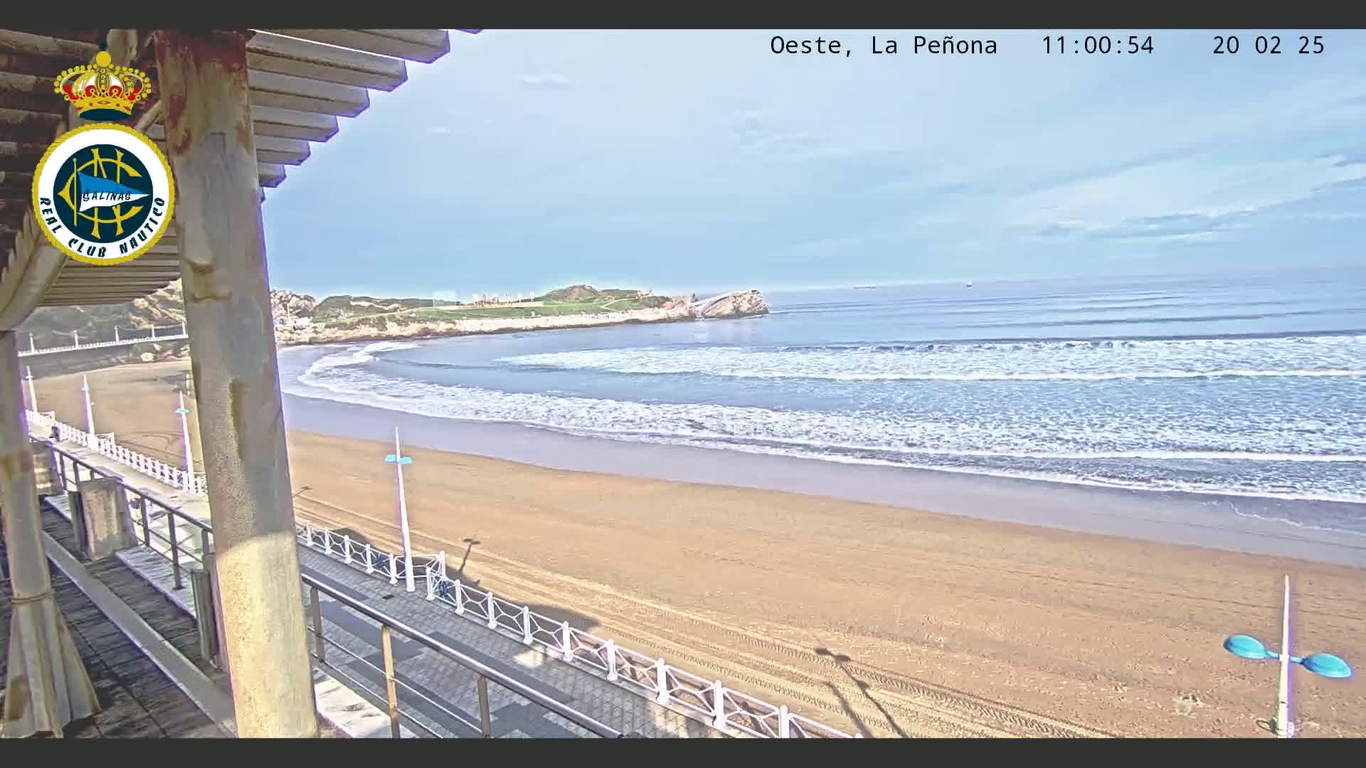 A sandy beach with gentle waves rolls onto the shore under a mostly clear sky, viewed from an elevated position on a boardwalk.
