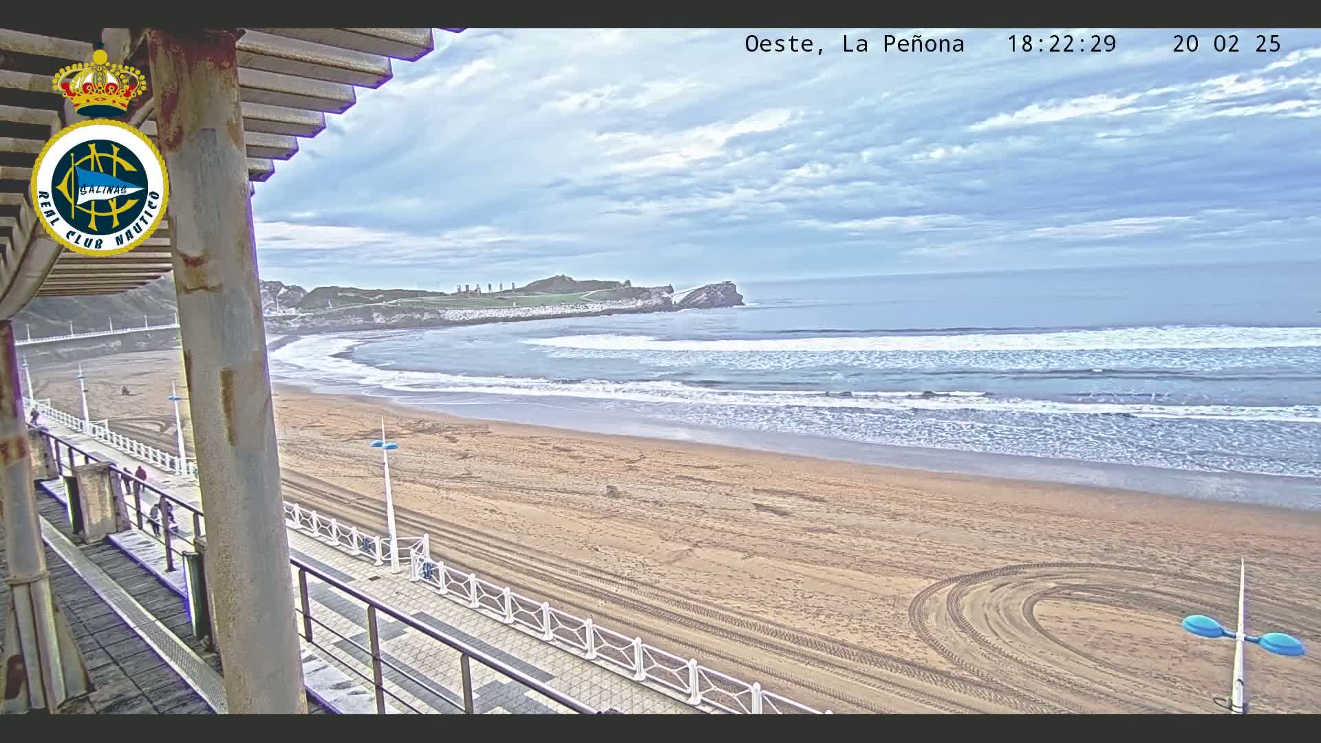 A sandy beach with gentle waves under a partly cloudy sky is viewed from a boardwalk overlooking the ocean.