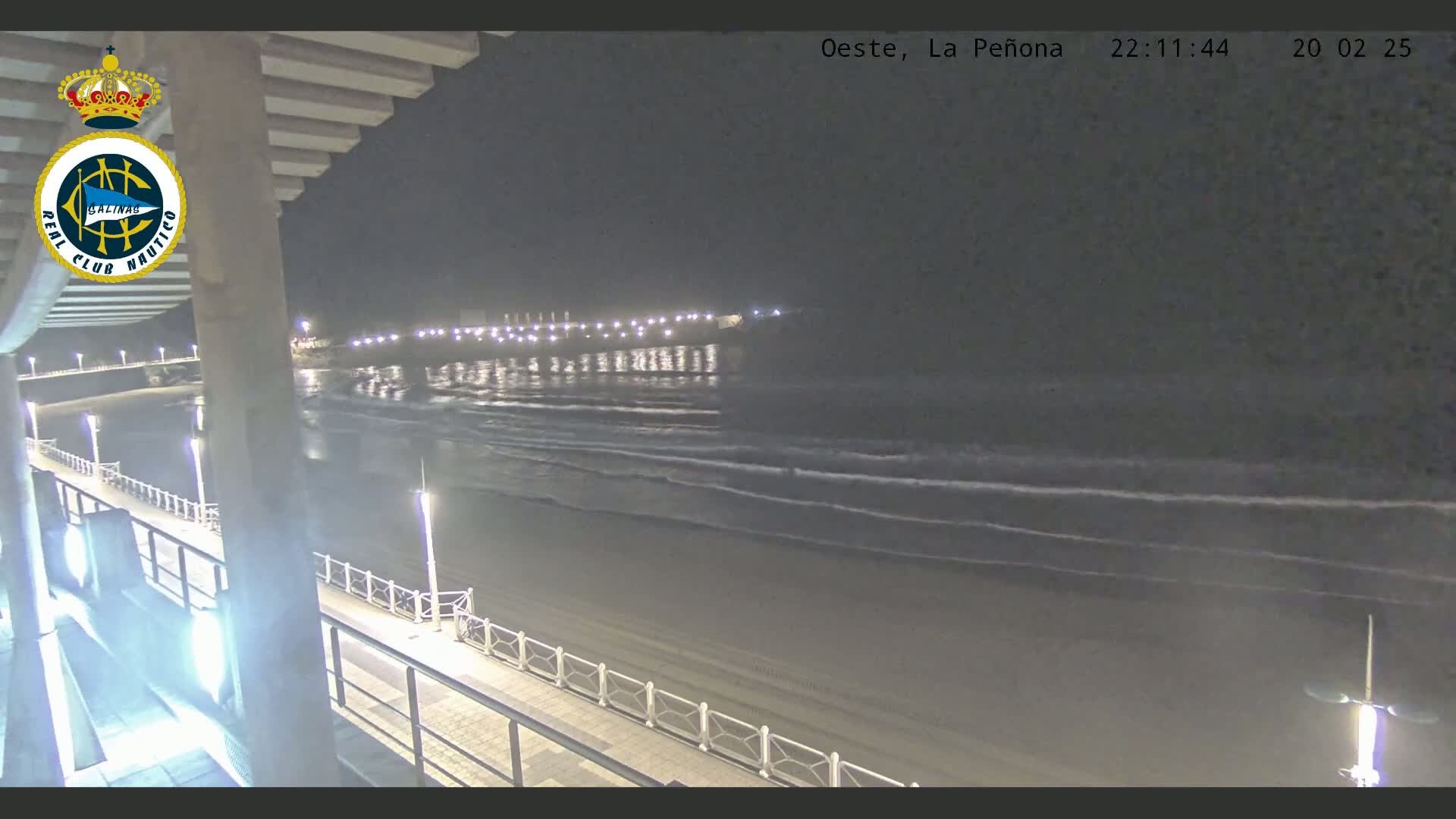 A nighttime view from an elevated position shows a mostly calm ocean with small waves lapping a sandy beach, illuminated by lights along a boardwalk.