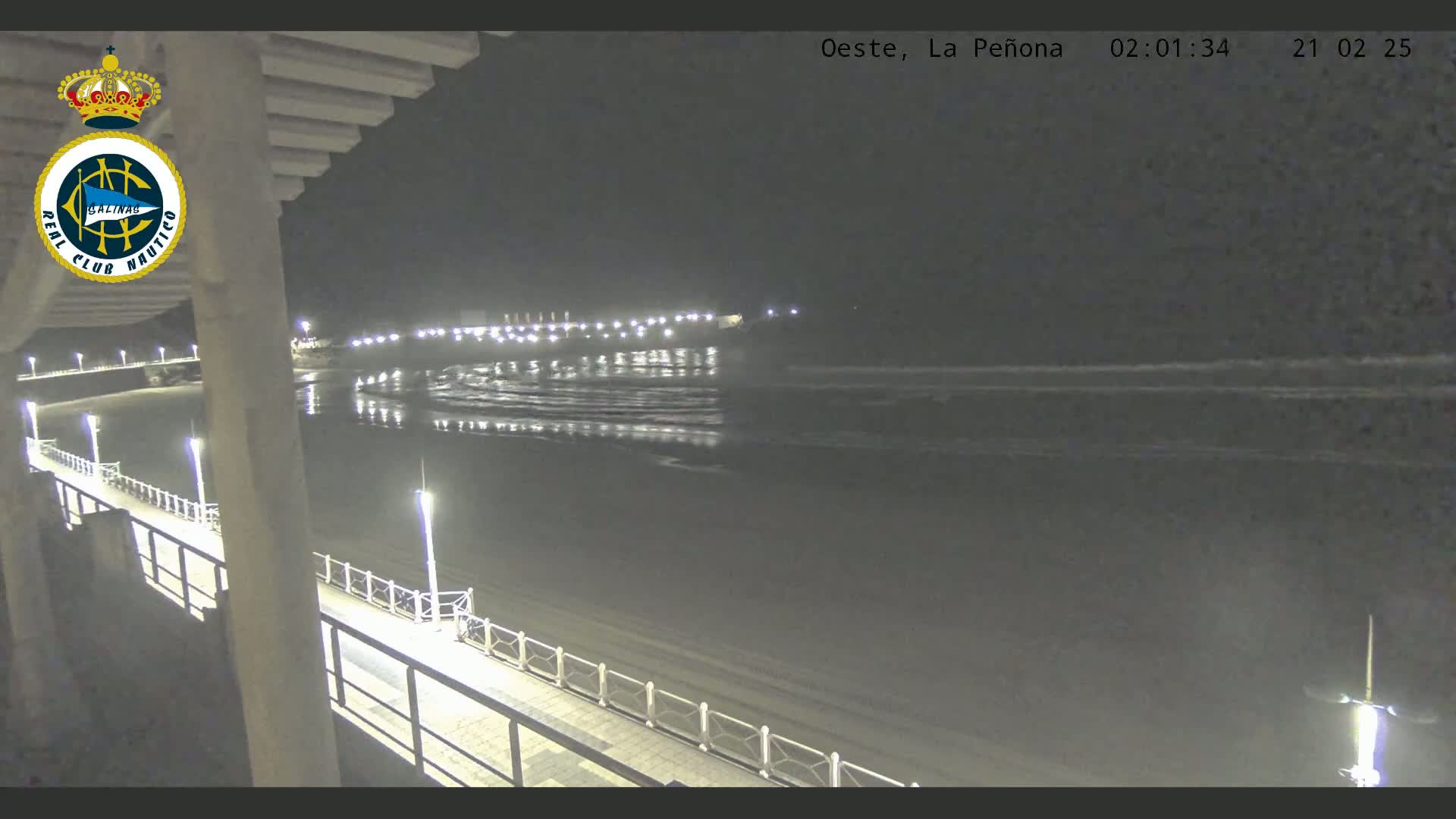 A nighttime view from a pier shows a calm, dark ocean reflecting lights from a distant shoreline.