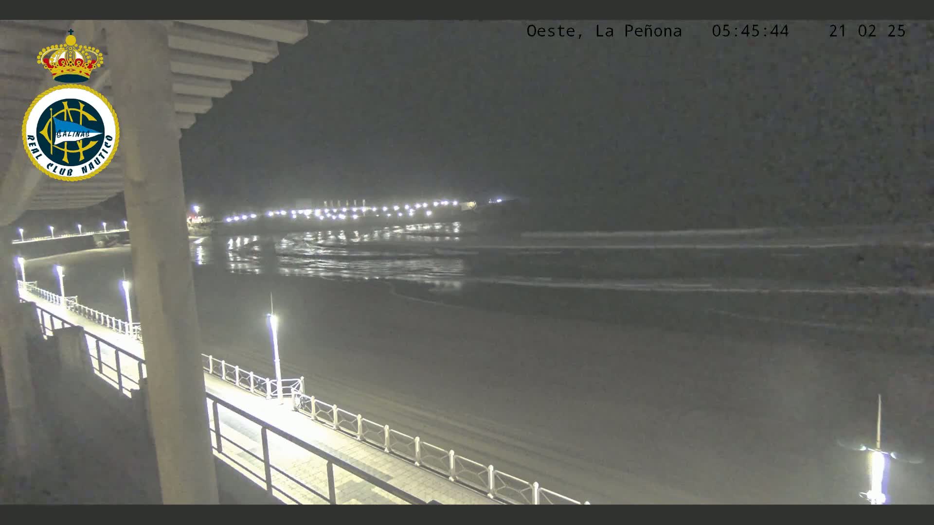 A nighttime view of a beach with gentle waves, a lighted boardwalk, and a distant shoreline with lights.