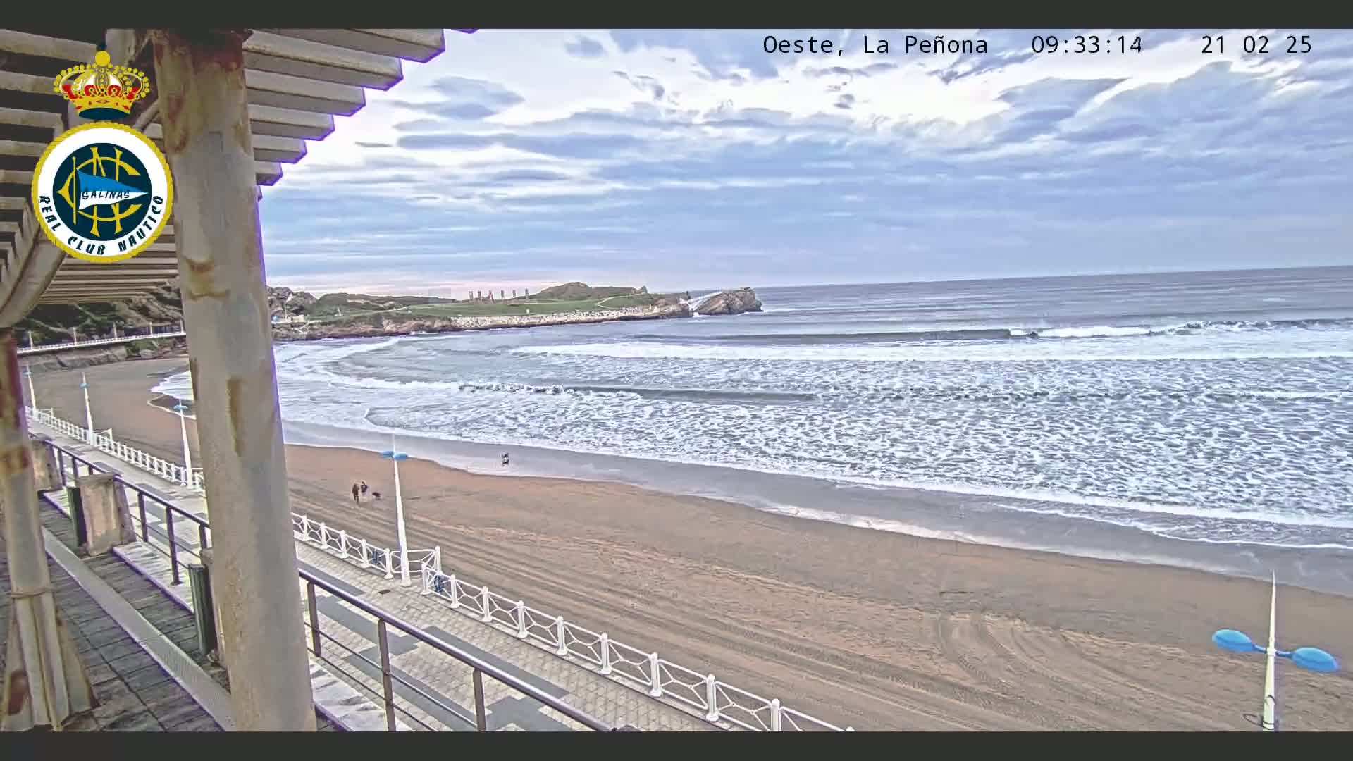 A sandy beach with gentle waves under a partly cloudy sky is viewed from a seaside structure.