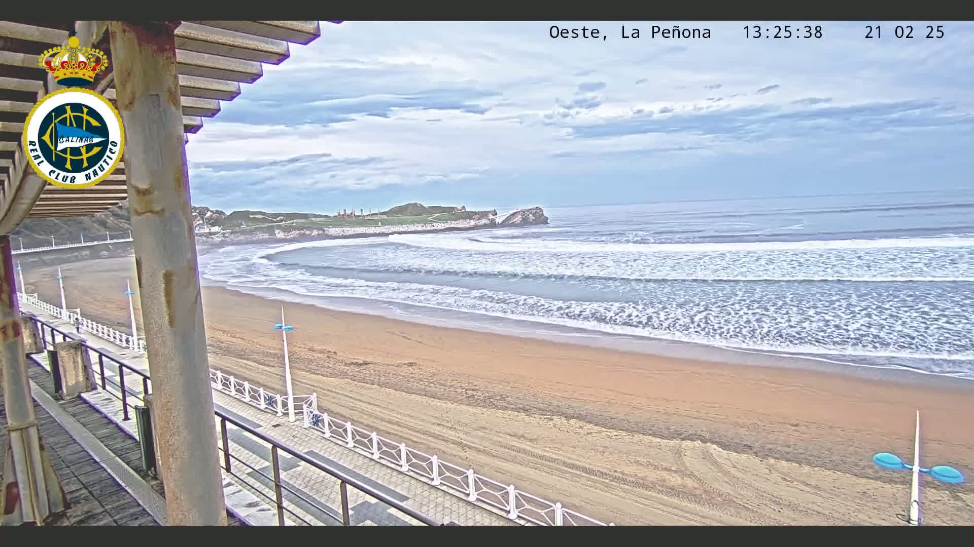A sandy beach with gently rolling waves under a partly cloudy sky is viewed from a boardwalk with a railing.