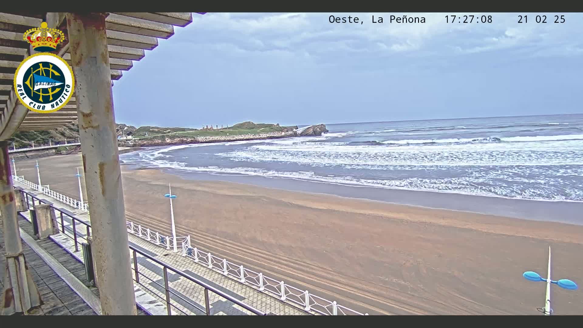A sandy beach with gentle waves under a partly cloudy sky is viewed from a raised vantage point with a railing and walkway.