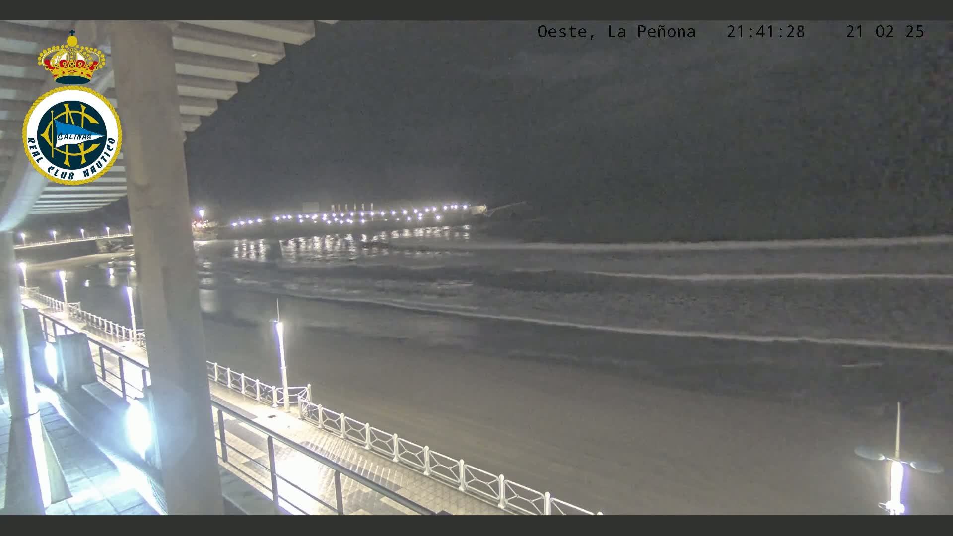 A nighttime view of a beach with gentle waves lapping the shore, illuminated by lights along a boardwalk and a pier in the distance.
