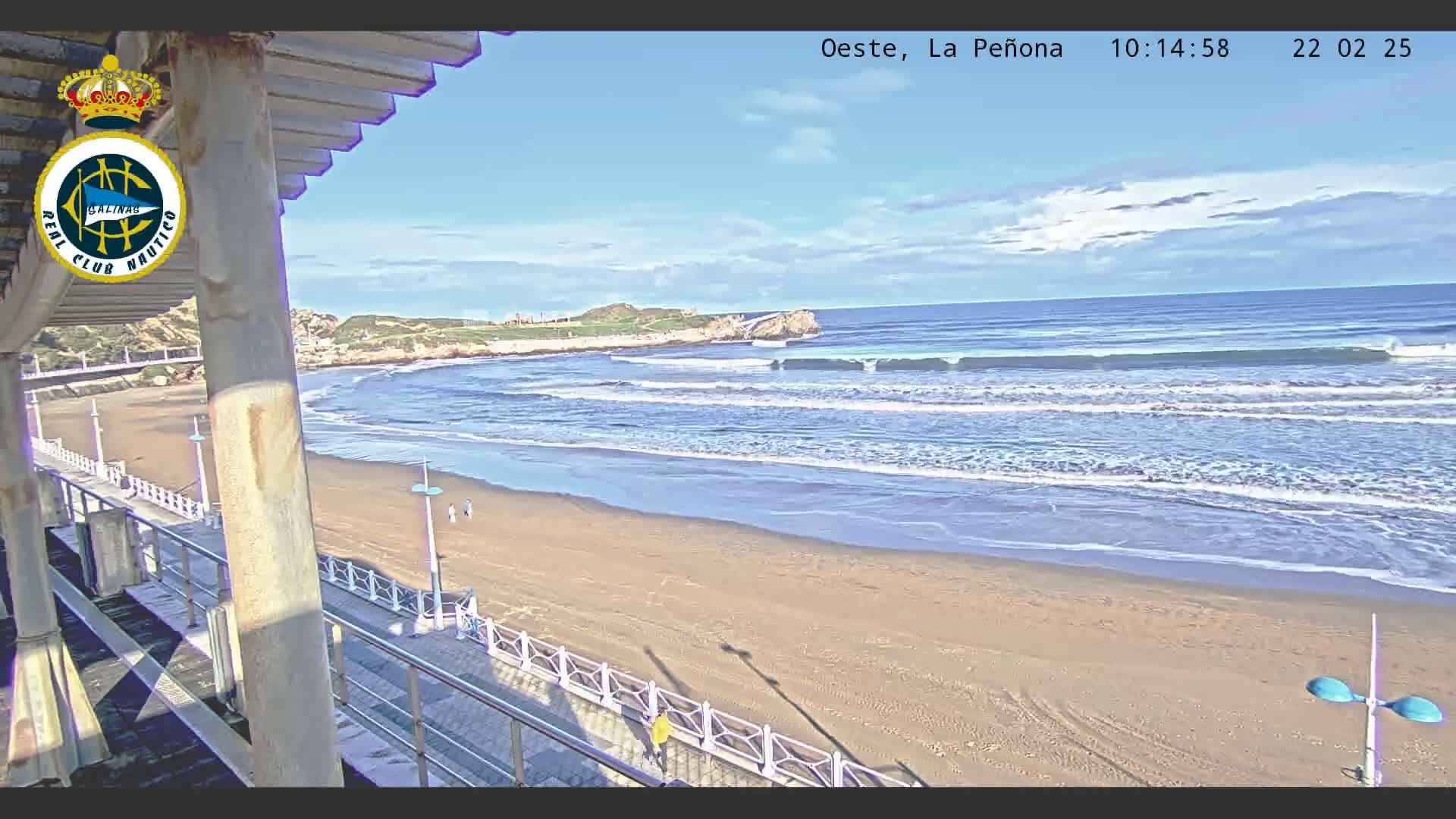 A sandy beach with gently rolling waves under a mostly sunny sky is viewed from a high vantage point, near a boardwalk and building.
