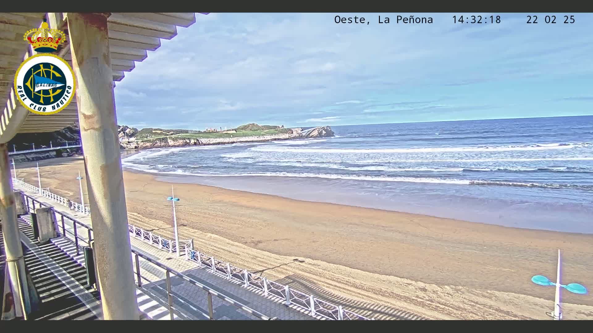 A sandy beach with gentle waves under a mostly sunny sky is viewed from an elevated vantage point, overlooking a boardwalk and a portion of a building.