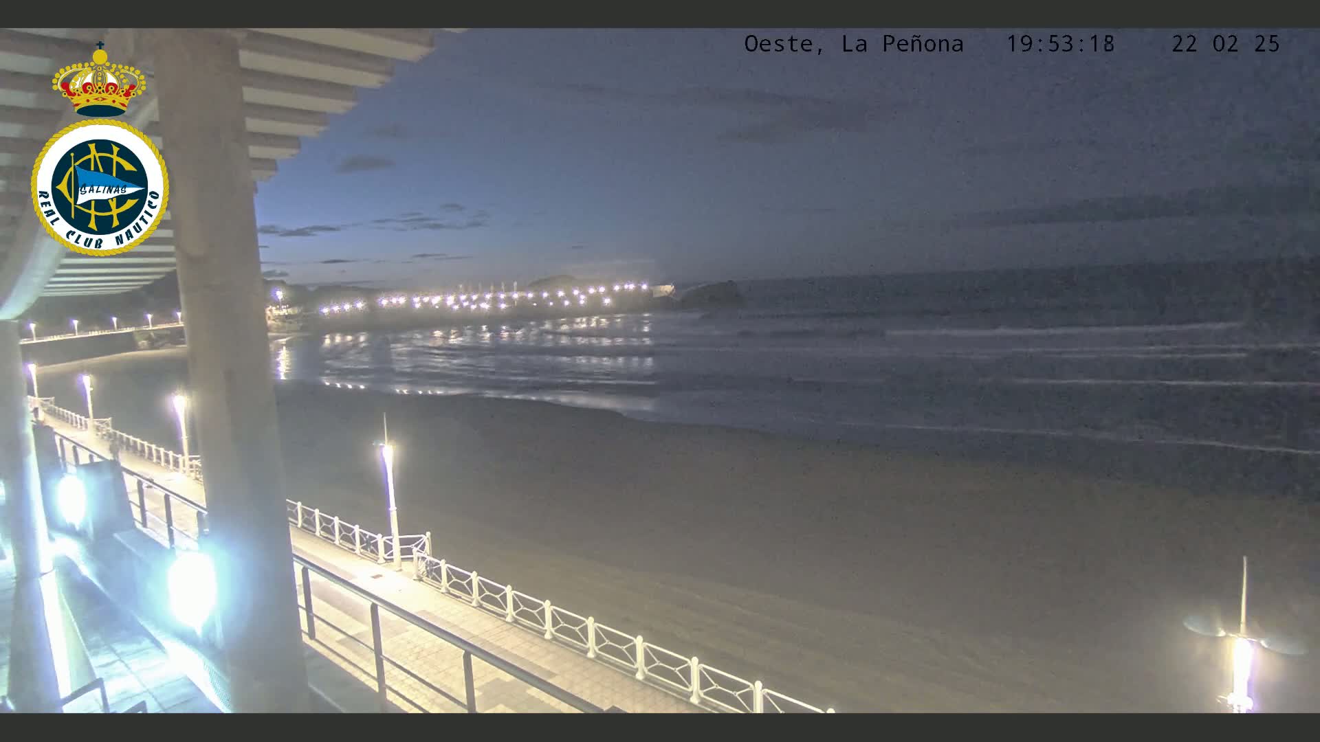 A nighttime view of a beach with calm waves, a boardwalk with lights, and a distant shoreline with illuminated buildings.