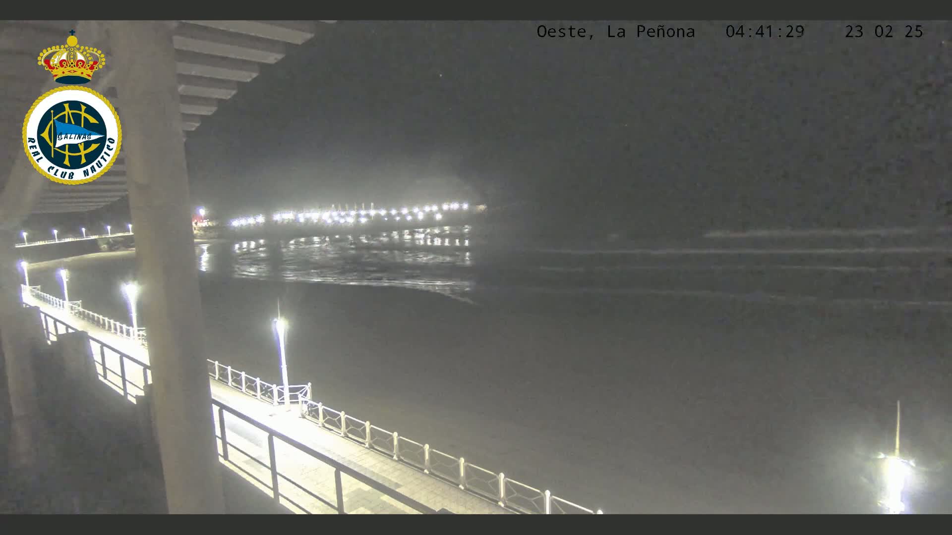 A nighttime view from a pier shows calm, dark water reflecting lights from a distant structure and the pier itself, under a clear sky.