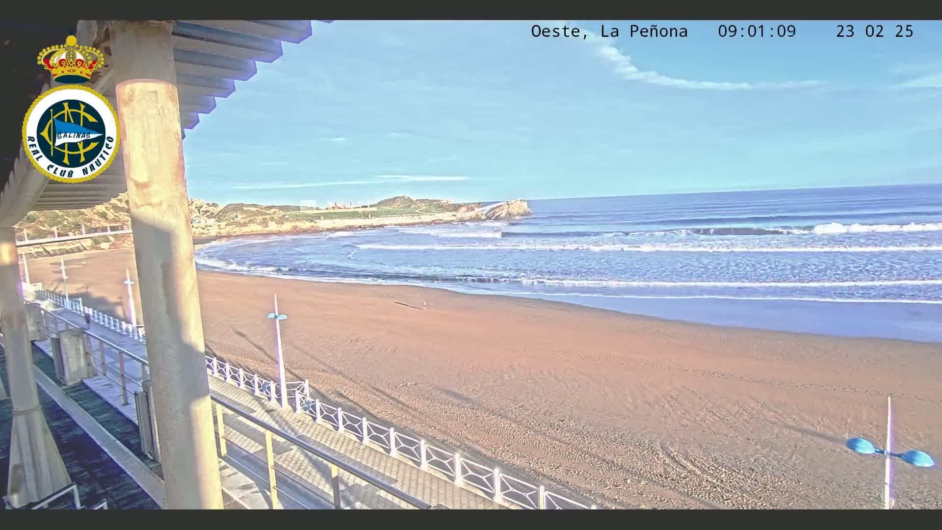 A sandy beach with gentle waves under a clear, sunny sky is viewed from an elevated position, overlooking a boardwalk and railing.