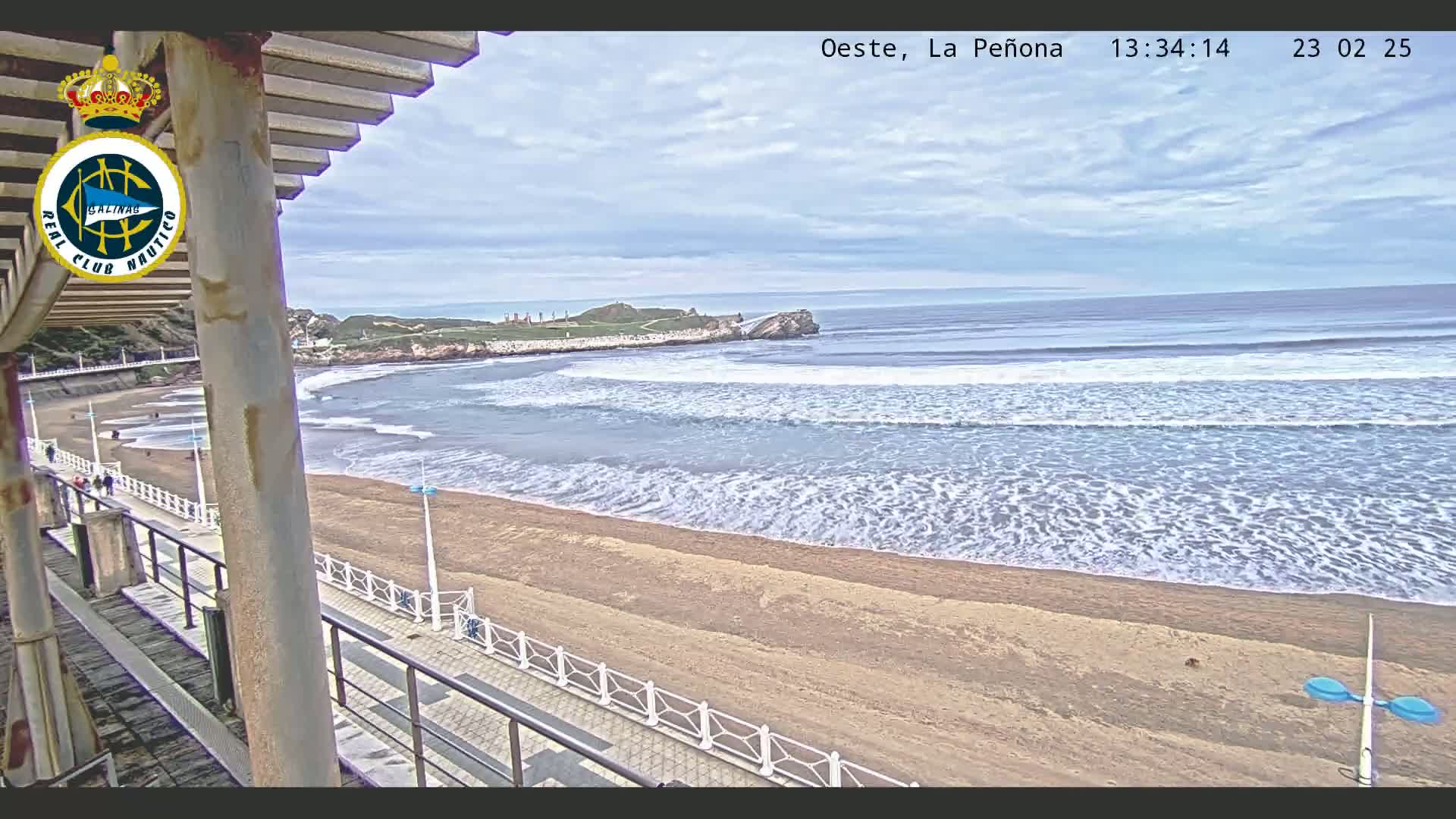 An overcast sky overlooks a sandy beach with gentle waves rolling onto the shore, viewed from an elevated position on a boardwalk with a railing.