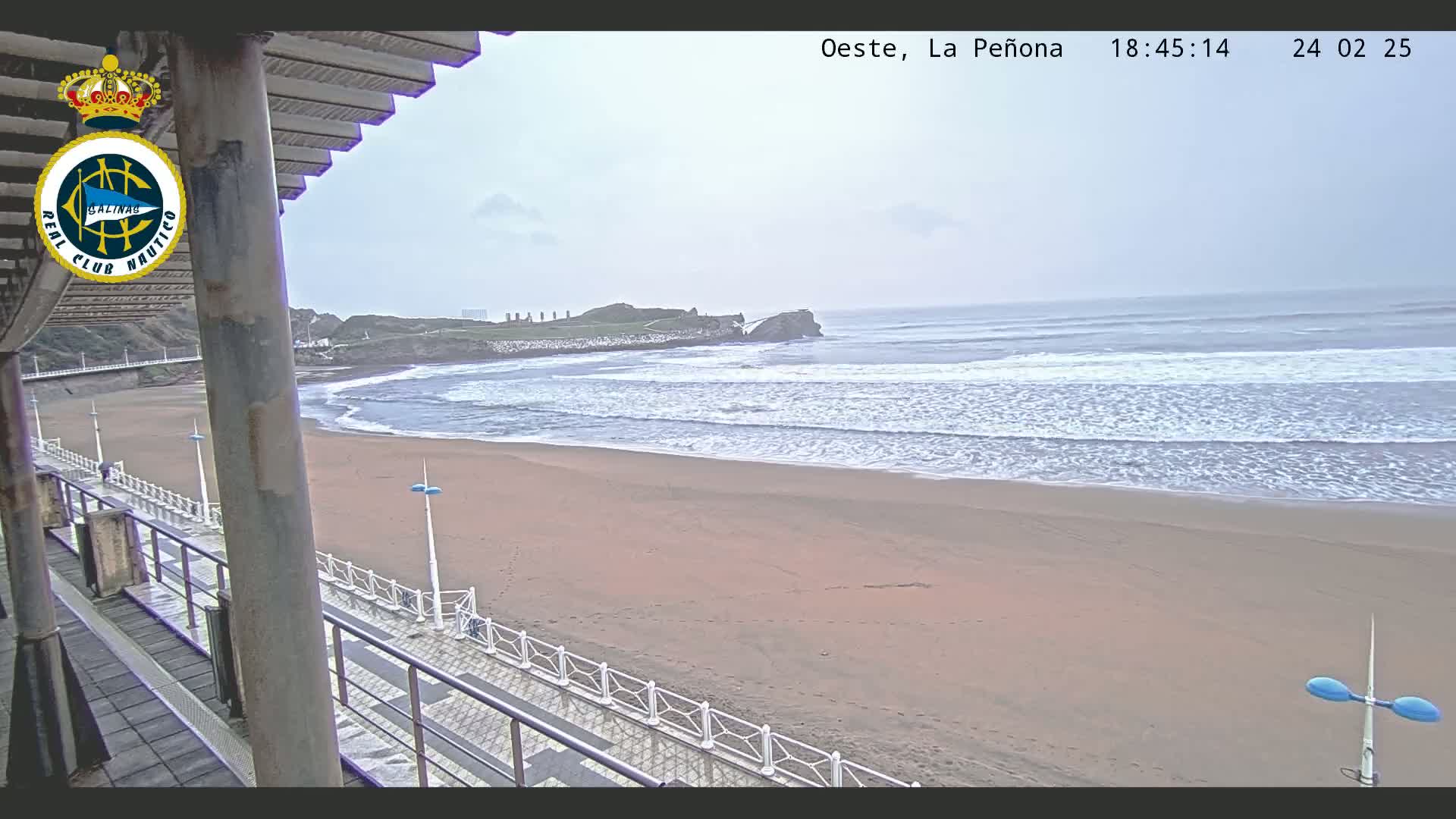 A sandy beach with gentle waves rolls onto the shore under an overcast sky, viewed from an elevated vantage point with a walkway and railing in the foreground.