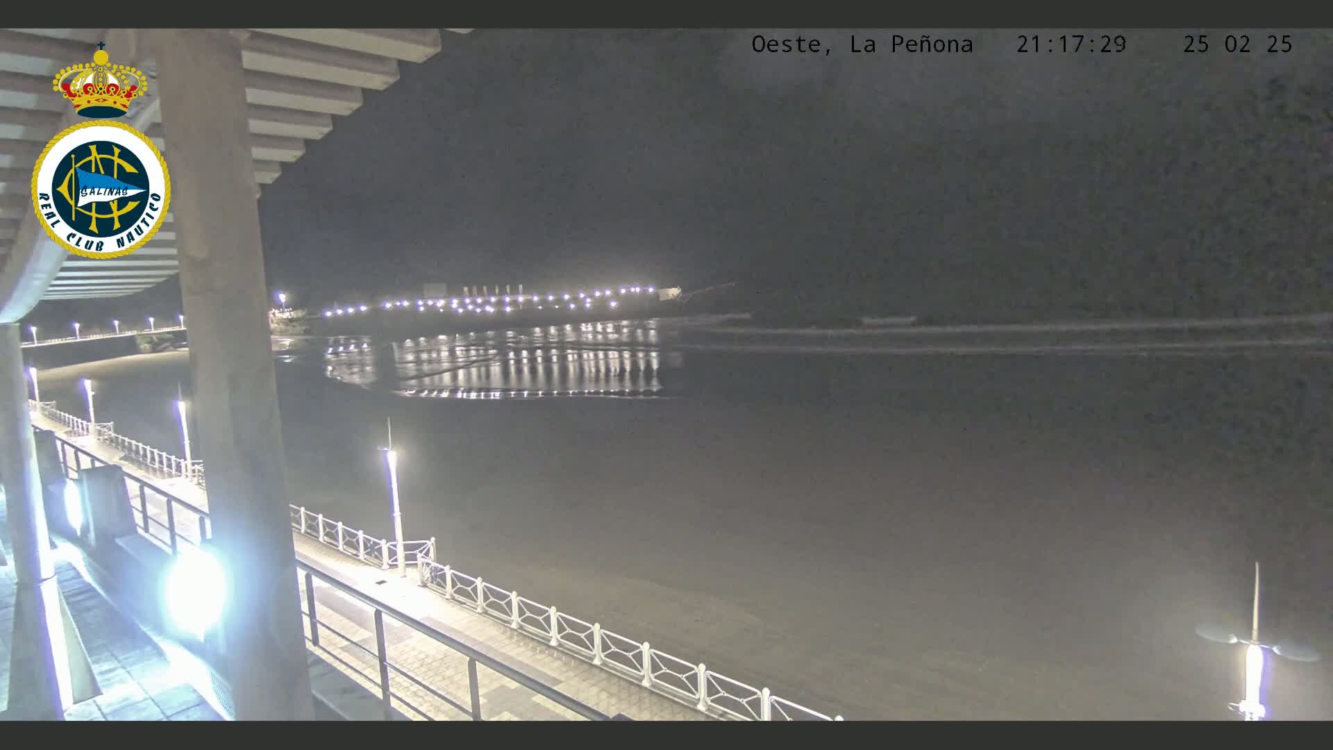 A nighttime view from a pier shows a calm body of water reflecting lights from a distant shore, under a dark sky.