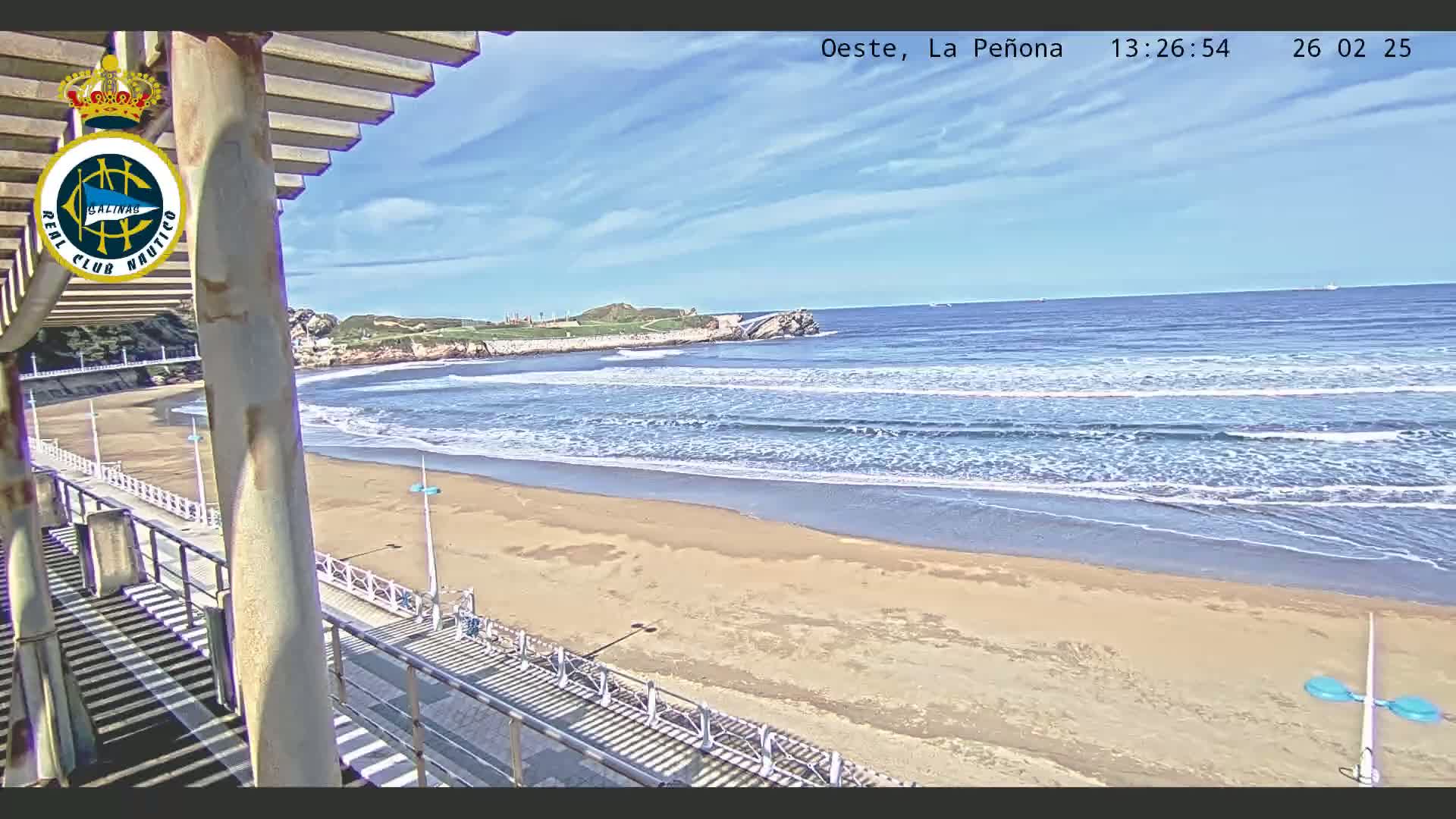 A sandy beach with gentle waves under a mostly sunny sky is viewed from an elevated position on a boardwalk.