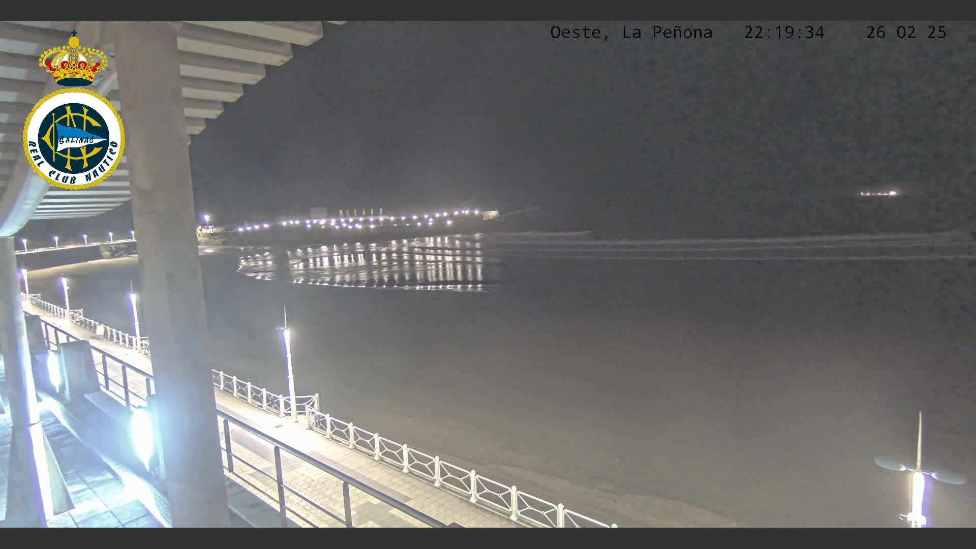 A nighttime view from an elevated position shows a dark beach, calm water reflecting lights from a distant structure, and a paved walkway with railings stretching along the shoreline.