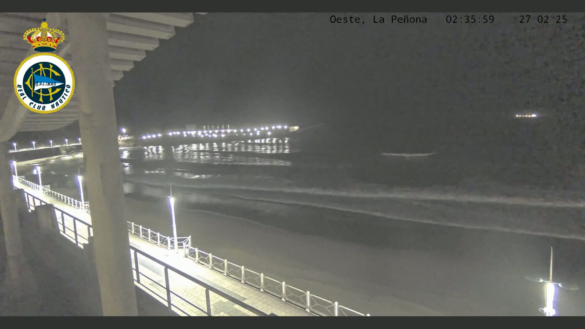 A nighttime view of a beach with gently lapping waves and a lit walkway along a pier.