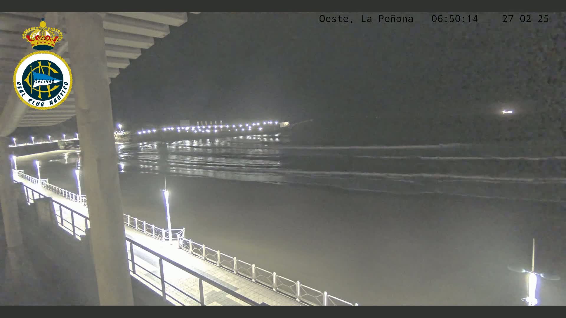 A nighttime view from a pier shows a dark ocean with gentle waves lapping the shore, illuminated by lights along the pier and a distant shoreline.