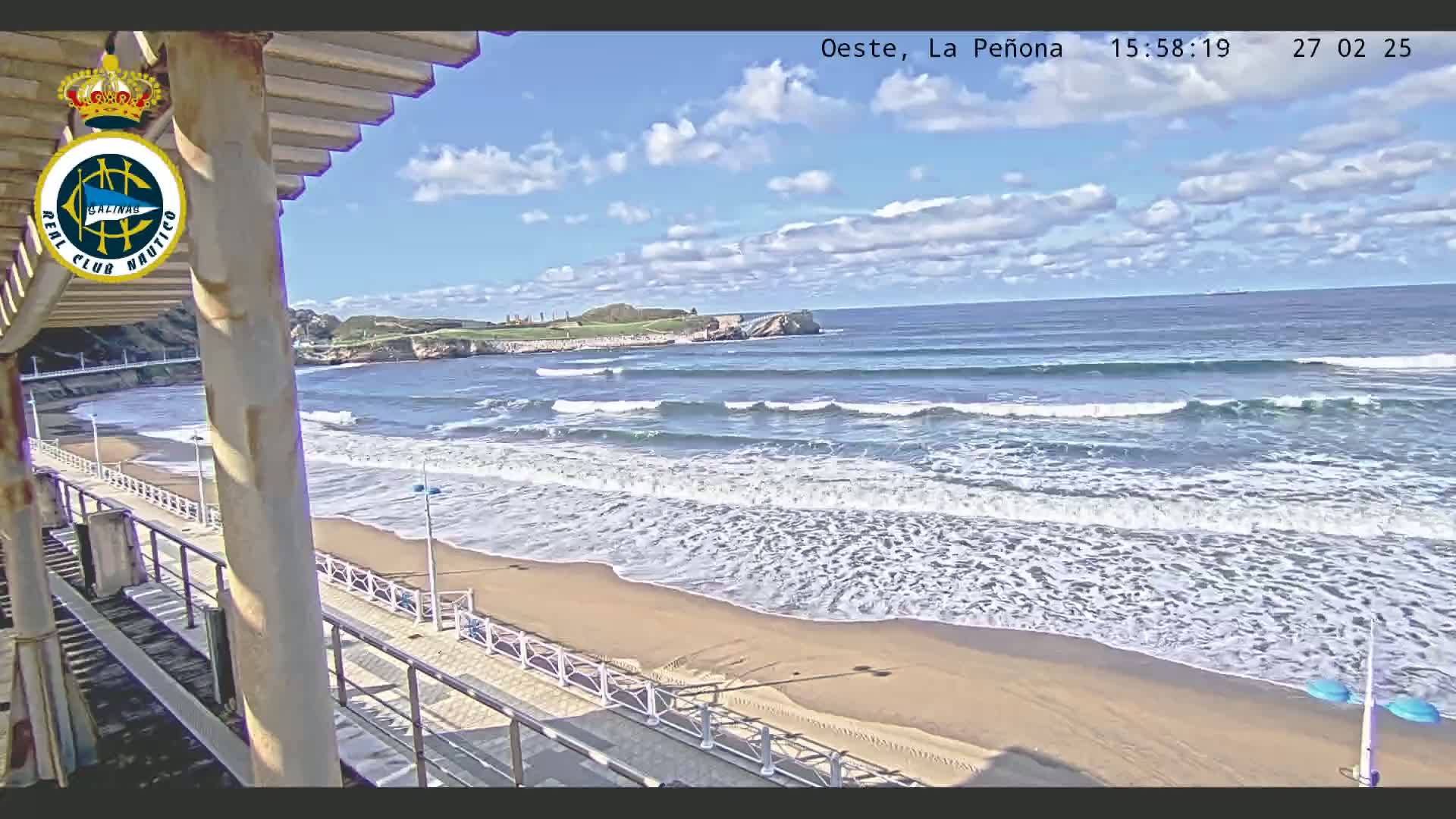A partly cloudy day shows a sandy beach with waves rolling onto the shore, viewed from a structure overlooking the ocean.