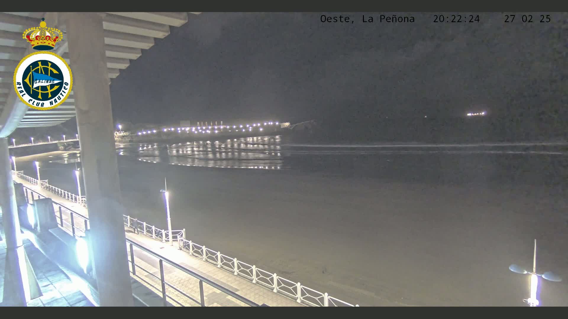 A nighttime view of a calm beach and ocean, with a walkway and lights visible along the shoreline.