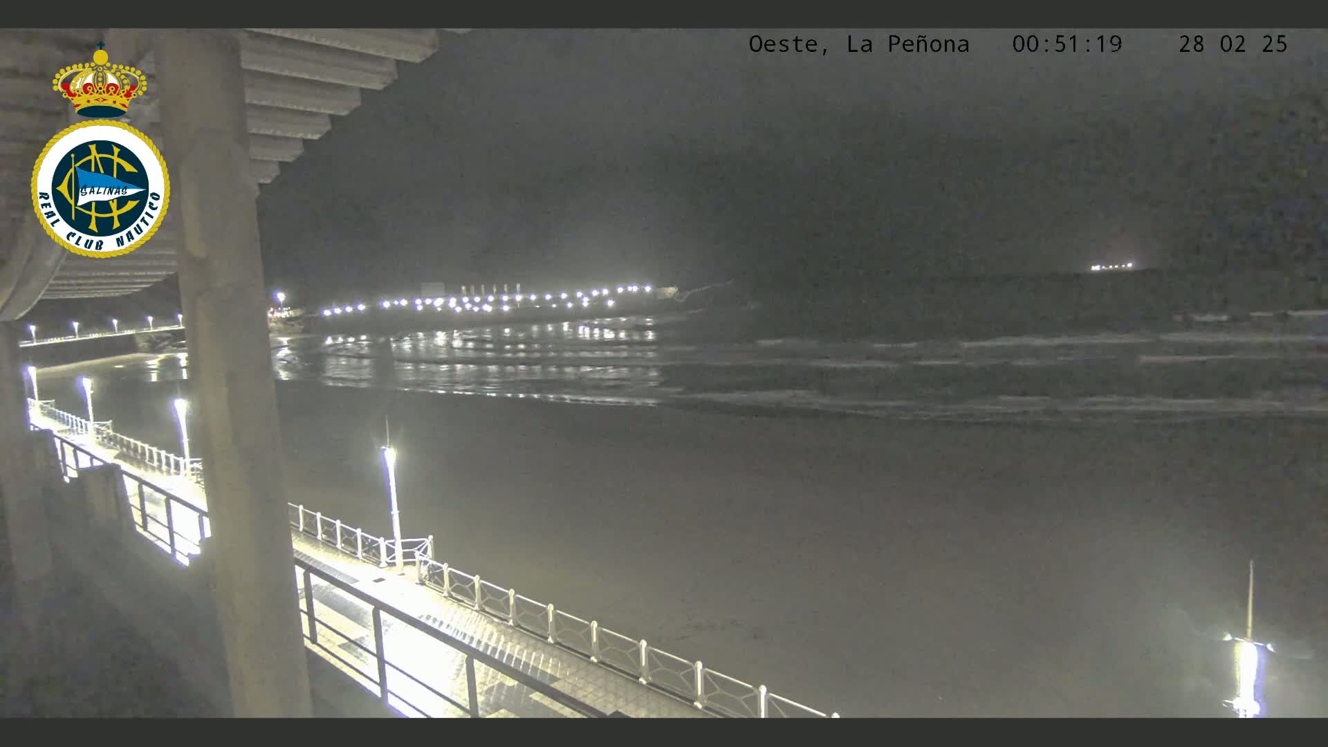A nighttime view from a pier shows a dark beach with gently rolling waves under a cloudy sky, illuminated by lights along the pier and in the distance.